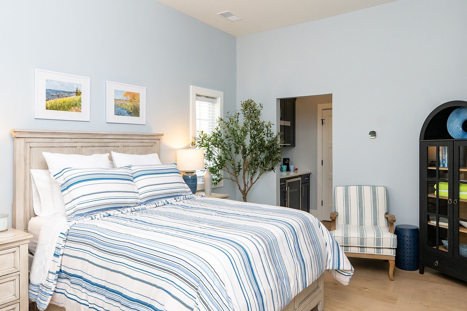 Bright bedroom with blue striped bedding, wood headboard, potted tree, striped armchair, and framed artwork on gray walls.