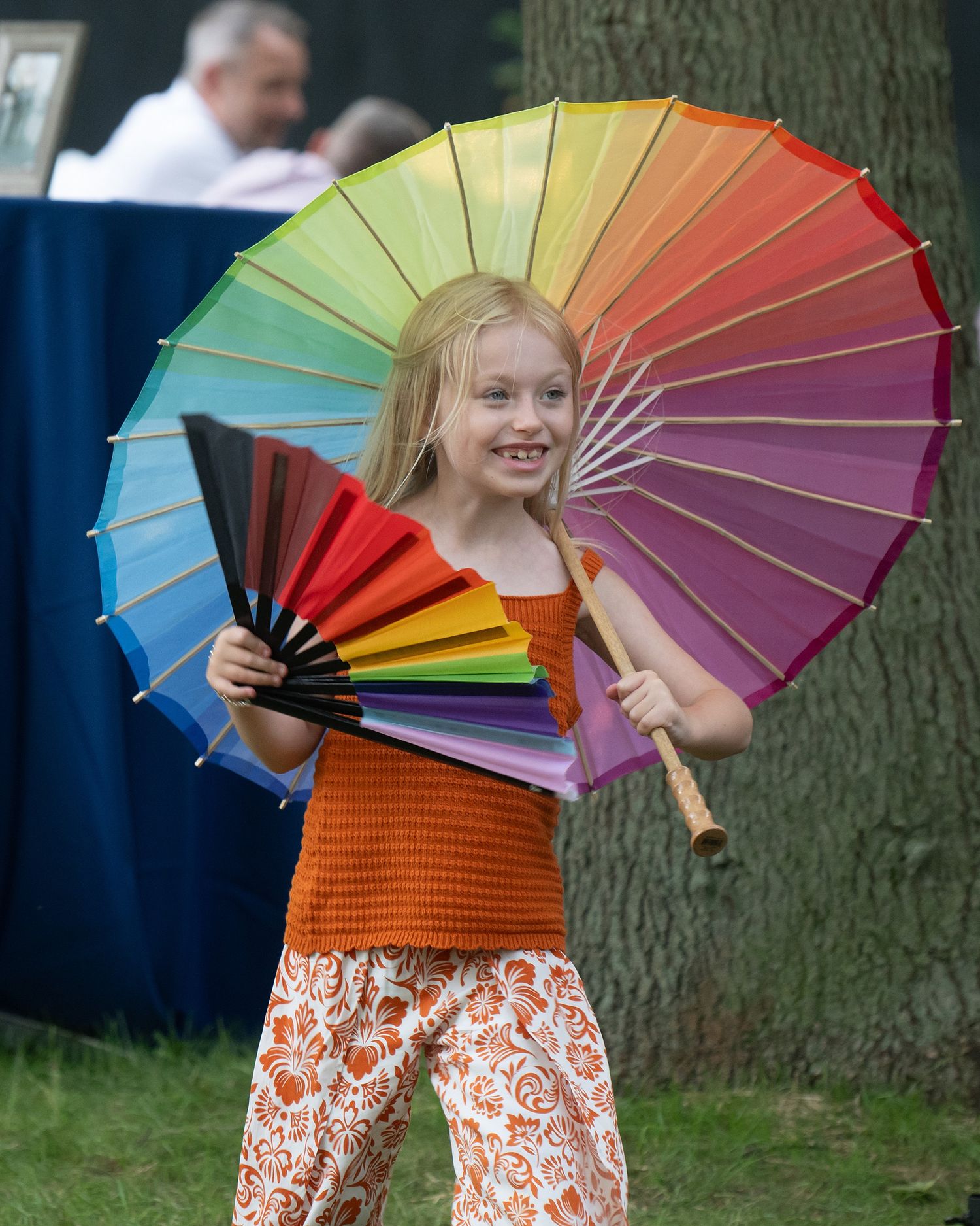 A colorful rainbow umbrella is held by a child wearing an orange top and floral pants at an outdoor event.