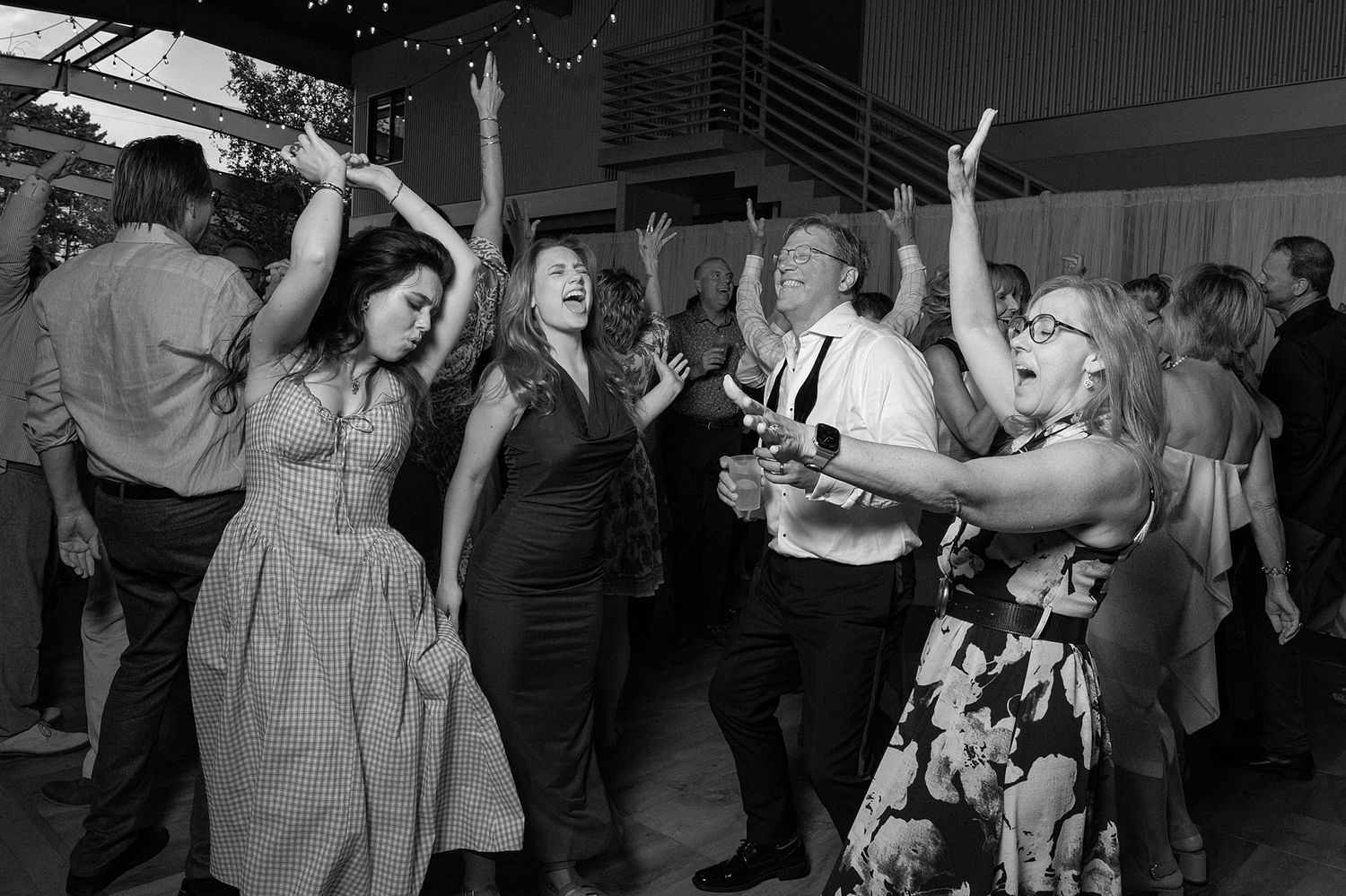 Black and white photo of guests dancing joyfully with arms raised at a lively wedding reception party.