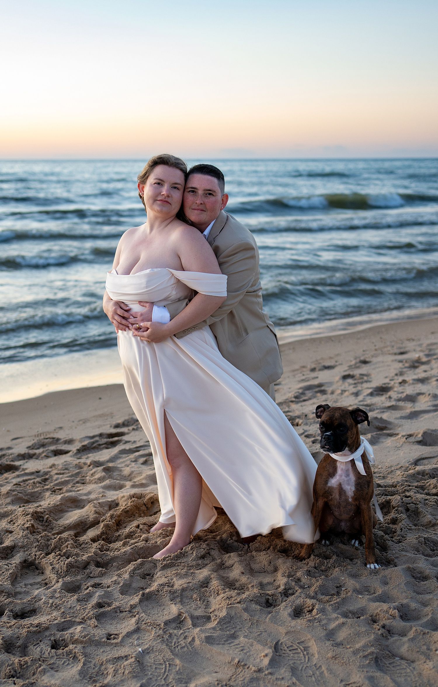 Couple in wedding attire embrace on a sandy beach at sunset with a brindle boxer dog wearing a white bow tie beside them.