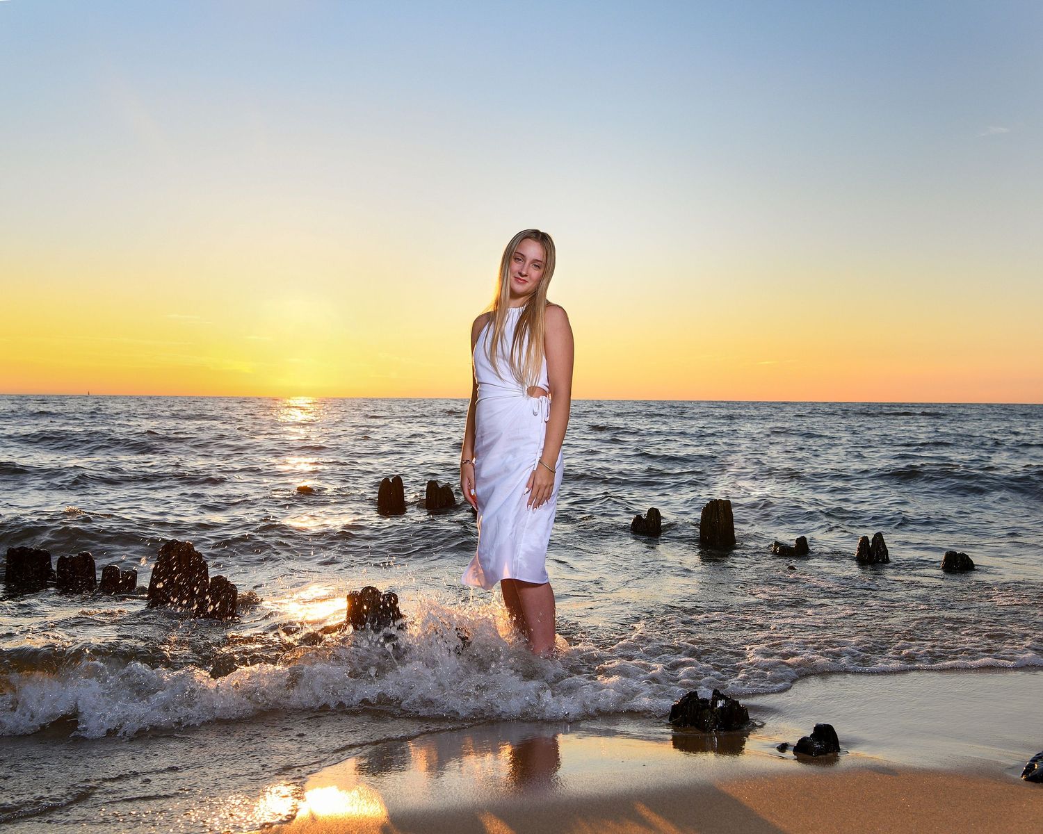 Figure in white dress walks along beach shore at sunset.