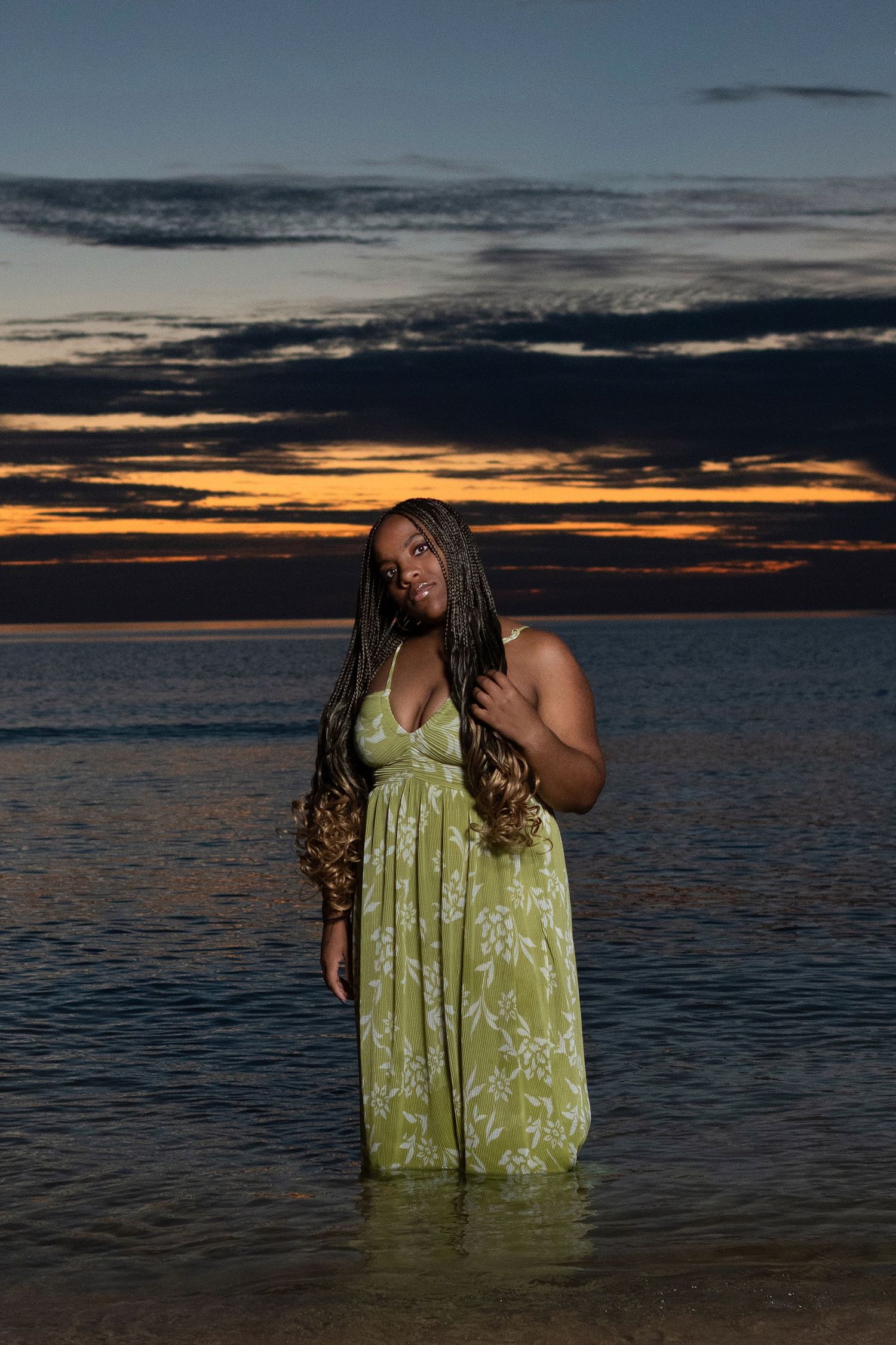 Person in lime green dress stands in shallow ocean water during dramatic sunset.