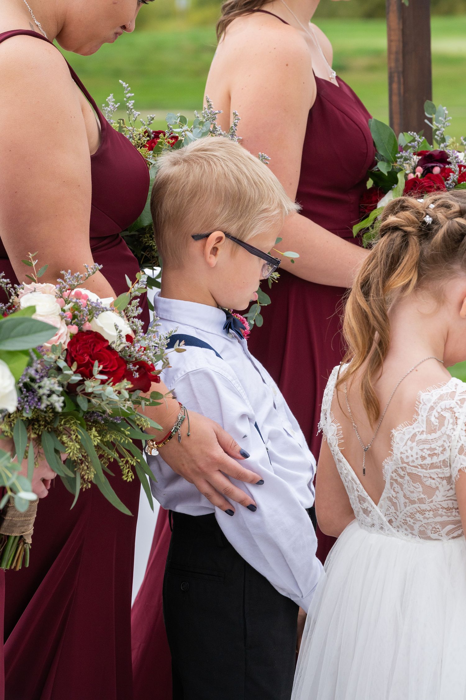 Young ring bearer in white shirt stands between bridesmaids in burgundy dresses during wedding ceremony.
