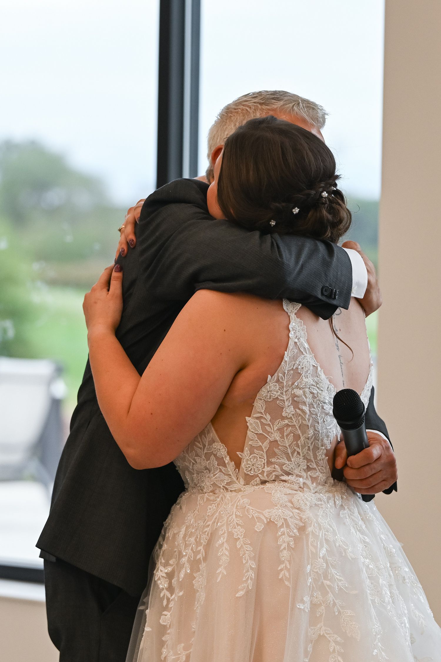Bride in lace gown embracing man in dark suit at wedding reception while holding microphone.