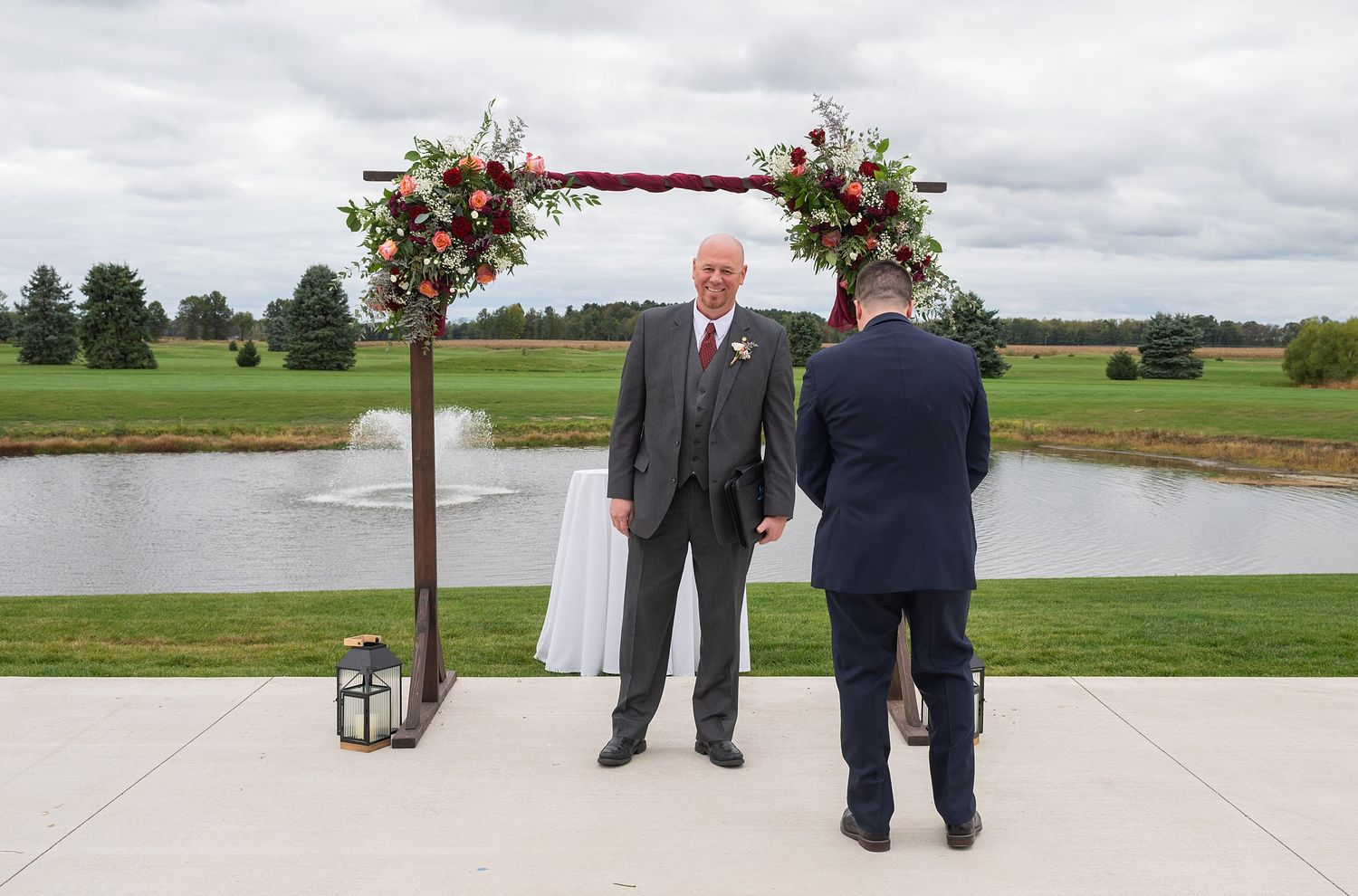 Groom stands under floral arch at outdoor wedding ceremony near pond, exchanging vows with officiant on cloudy day.