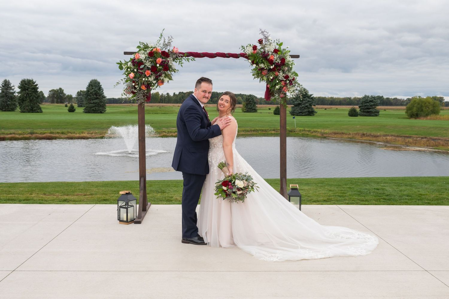 Bride and groom pose together under a rustic floral arch at an elegant outdoor wedding by a pond.