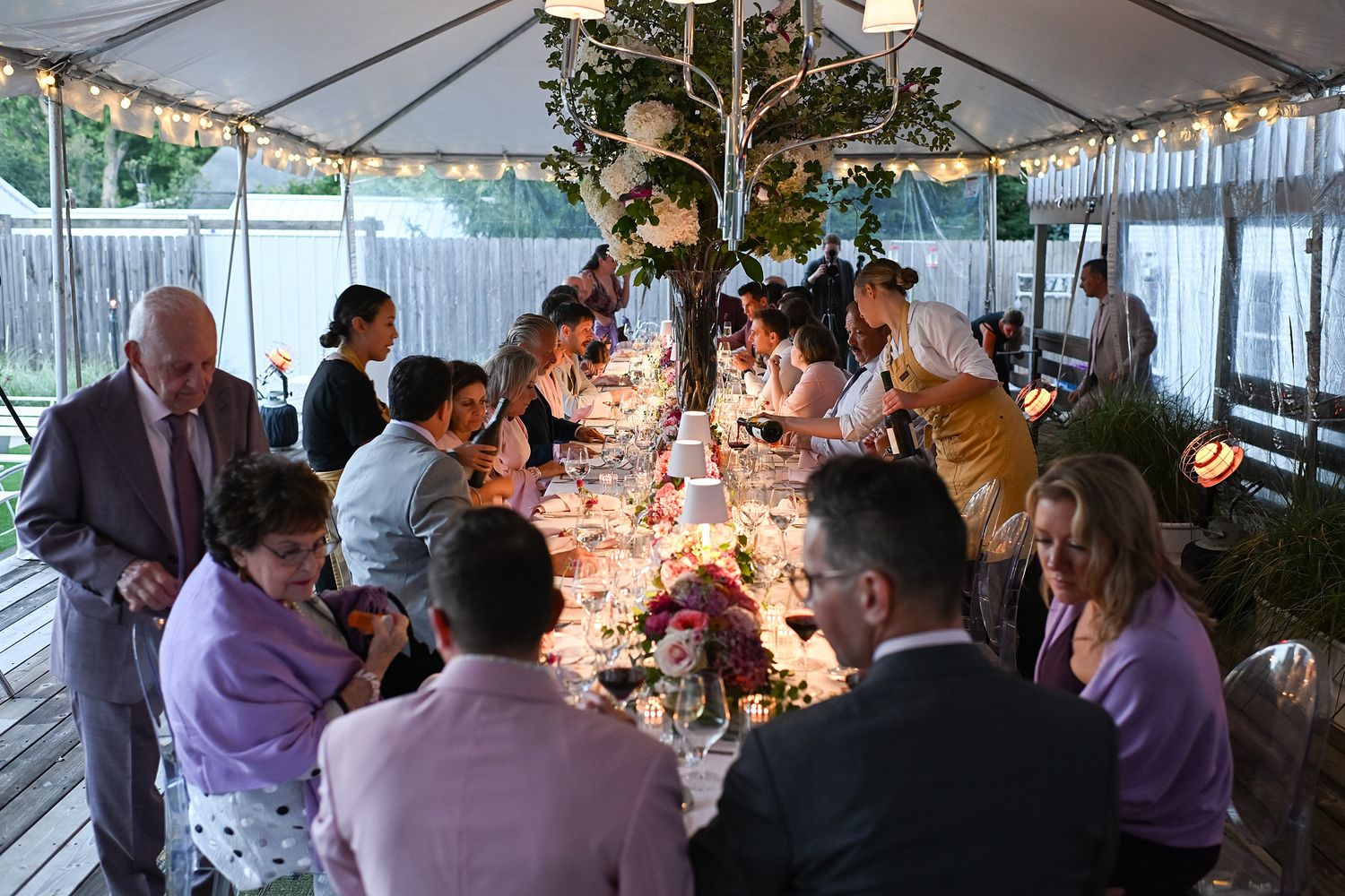 Guests gather under a white tent at an elegant evening wedding reception with glowing table centerpieces and string lights.