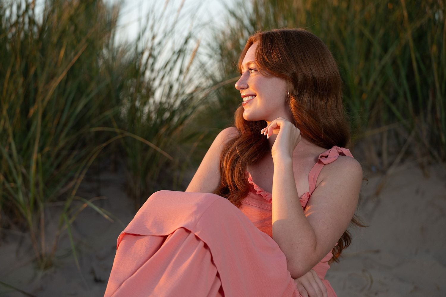 A smiling young woman with red hair sits on sandy beach dunes wearing a pink dress in warm golden sunlight.