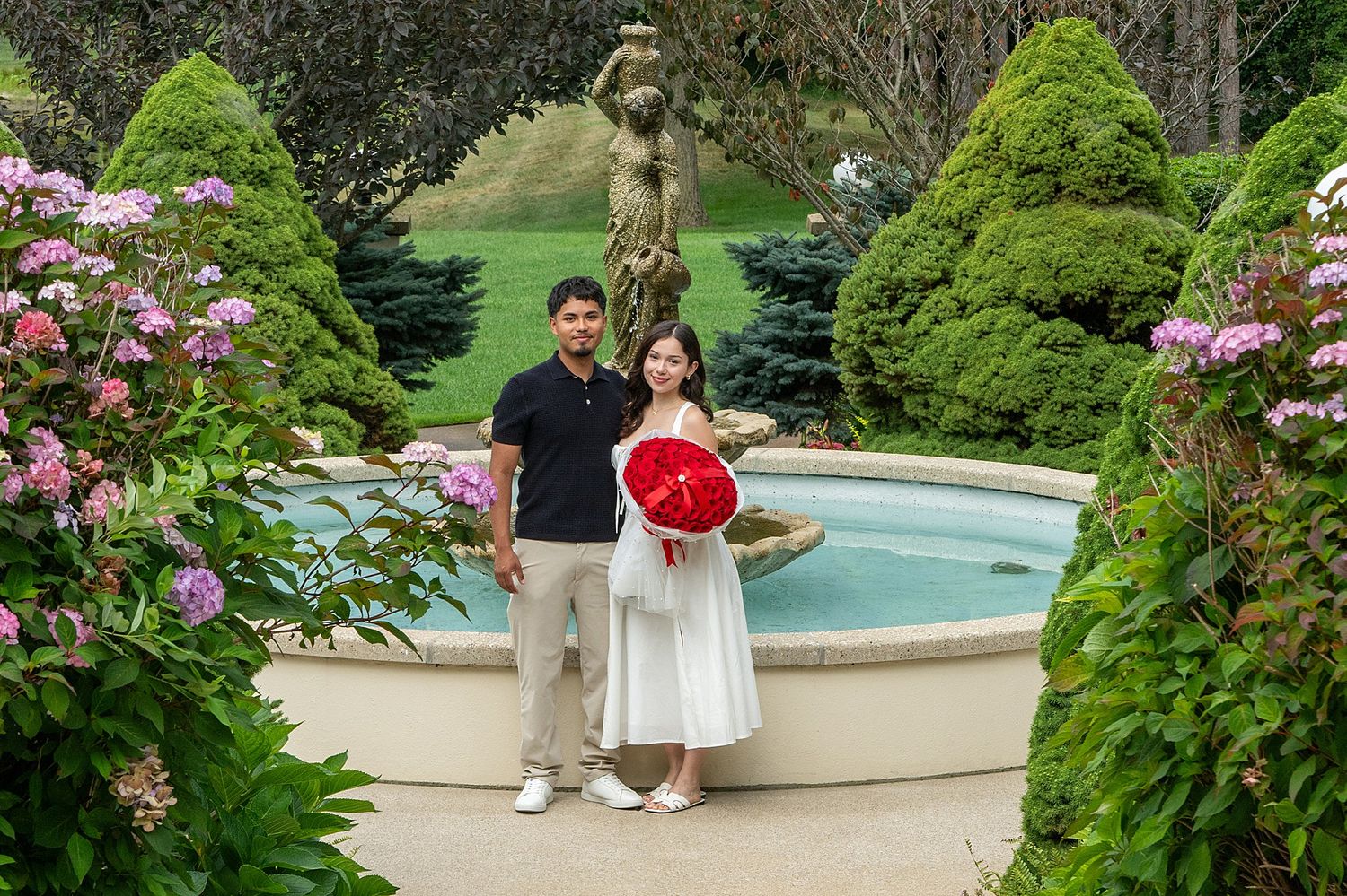 Couple posing by a garden fountain with lush greenery, pink flowers, and a red parasol.