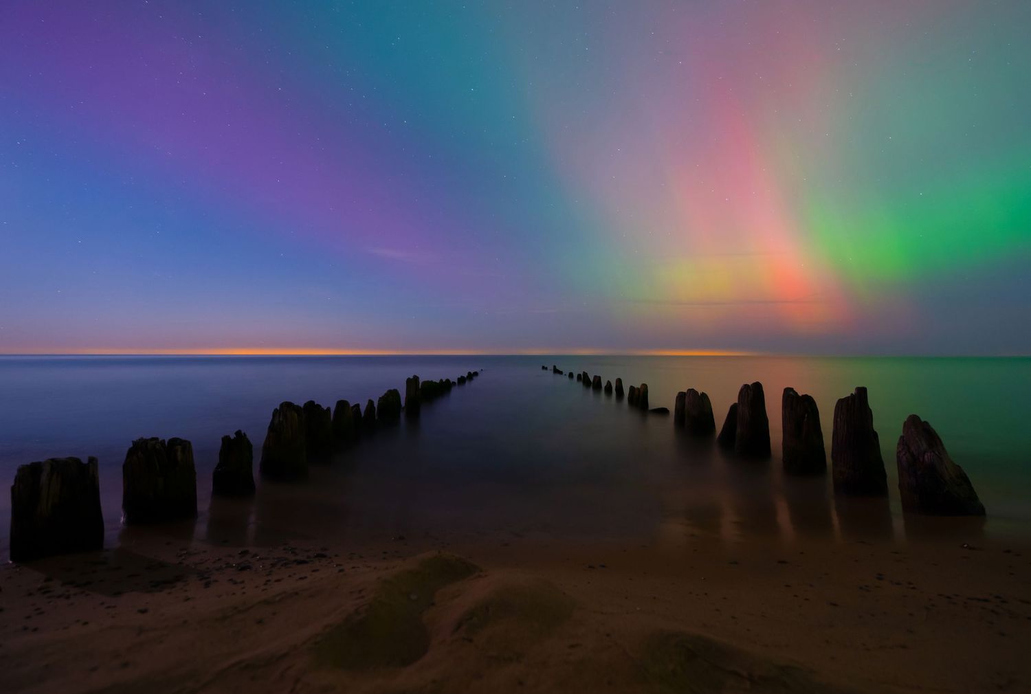 Aurora borealis illuminates colorful sky over calm sea with rows of weathered wooden posts on sandy beach.