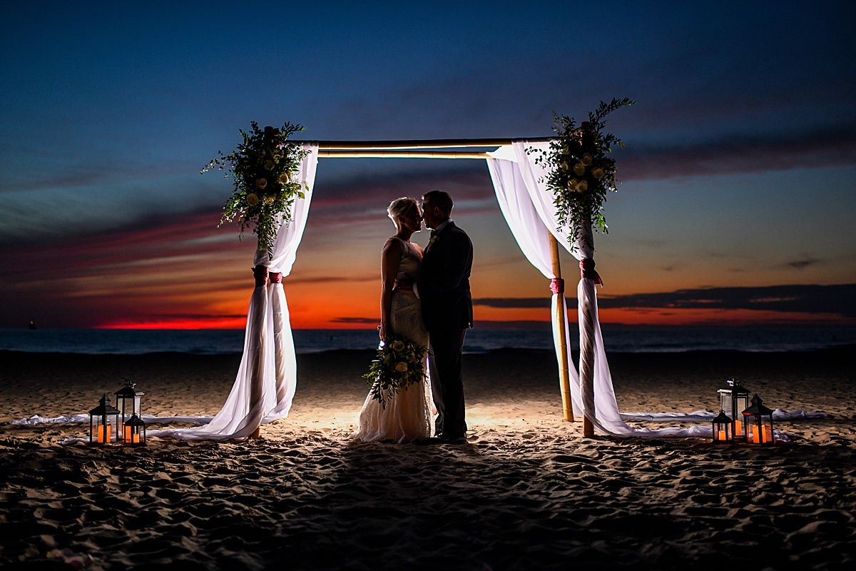 Couple kissing under floral arch at beach wedding during stunning sunset with candle lanterns on sand.
