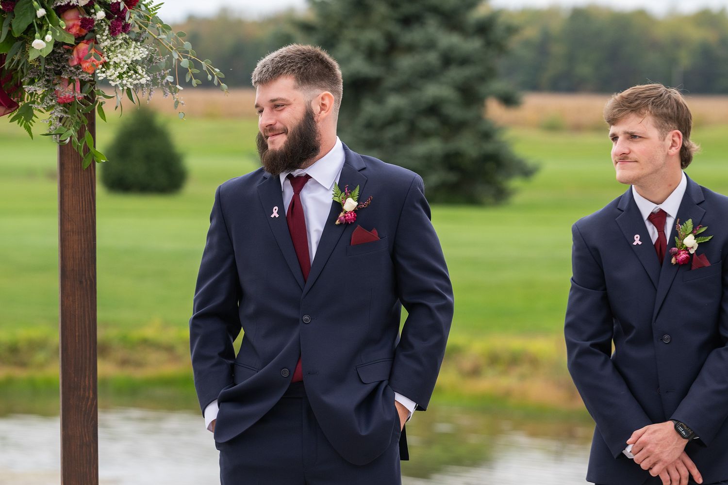 Two groomsmen in navy suits with burgundy ties stand at outdoor wedding altar.