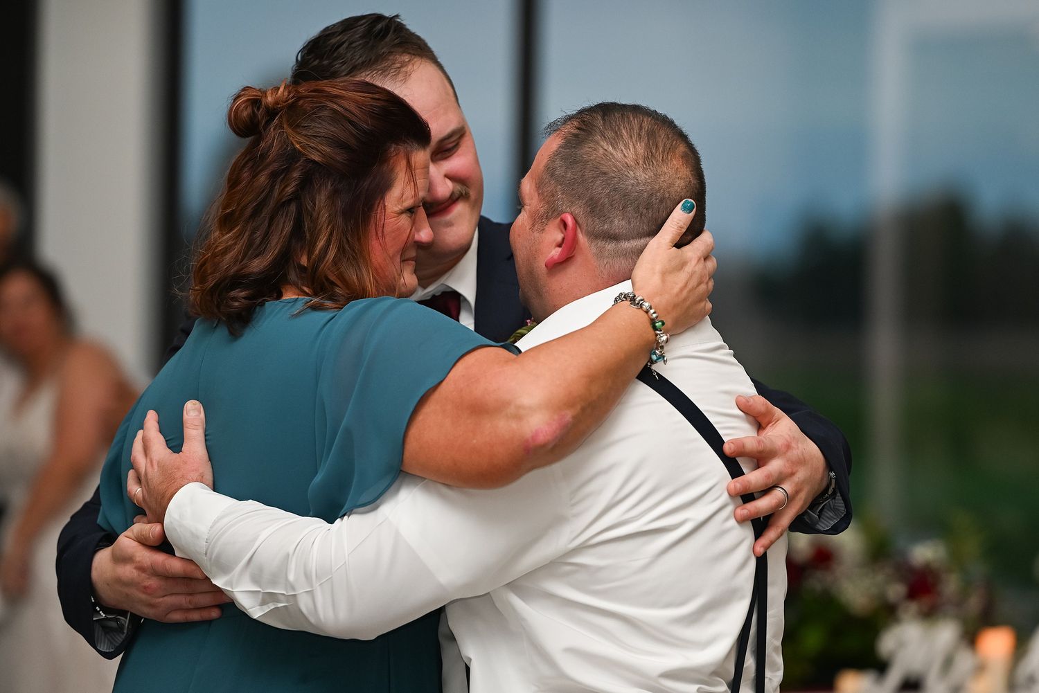 Emotional group hug between groom, mother in teal dress, and father at wedding reception celebration.