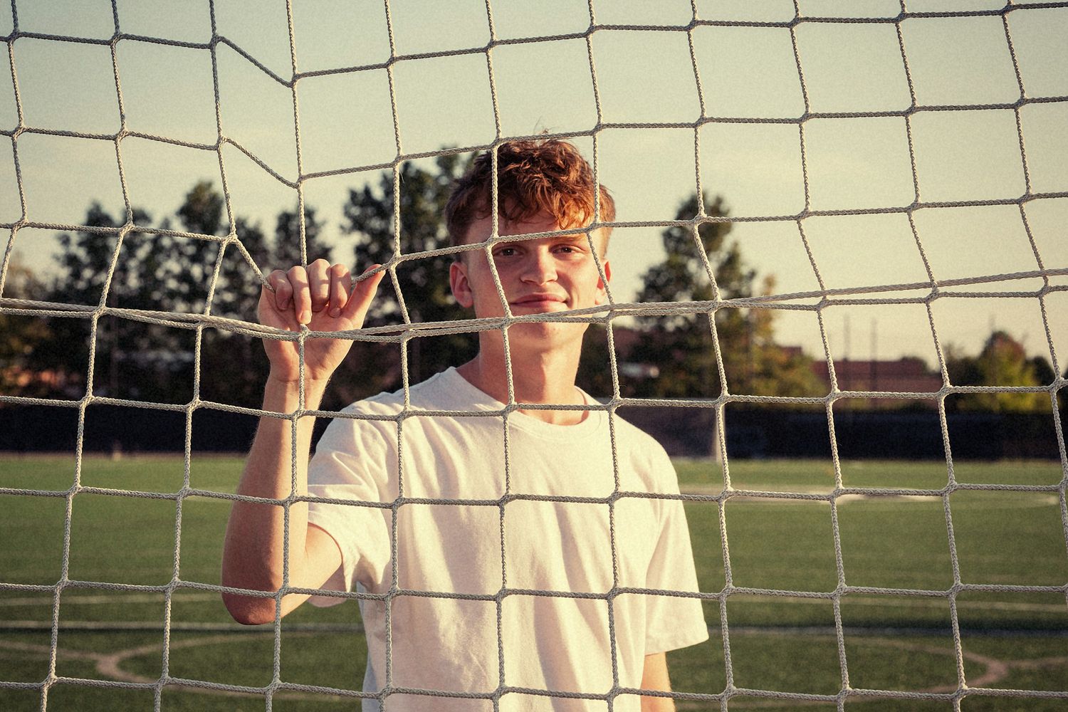 A soccer player poses behind a goal net during golden hour lighting.