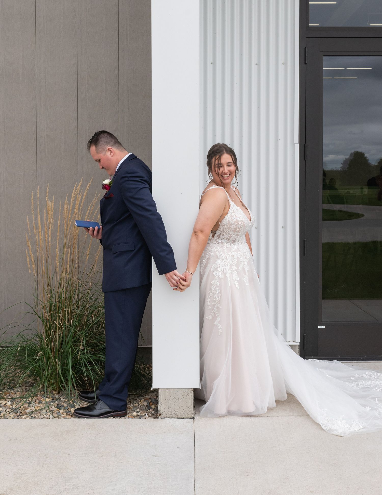 Bride in lace gown and groom in navy suit hold hands around a white pillar during a first look moment.