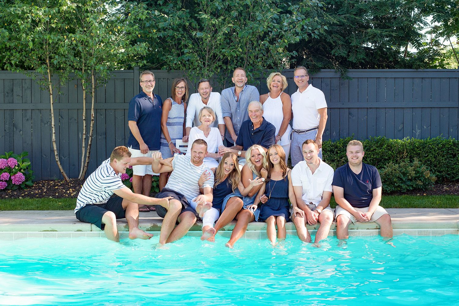 A group of friends sit along the edge of a swimming pool with their feet in the water on a sunny summer day.