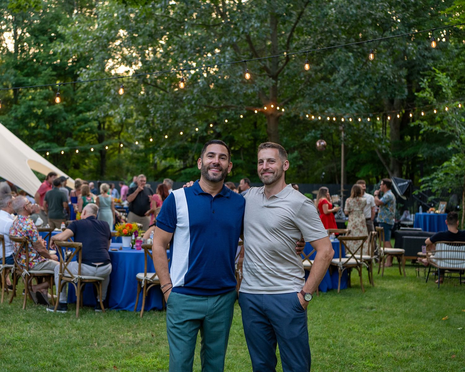 People enjoying an outdoor summer party with string lights and picnic tables in a lush garden setting.