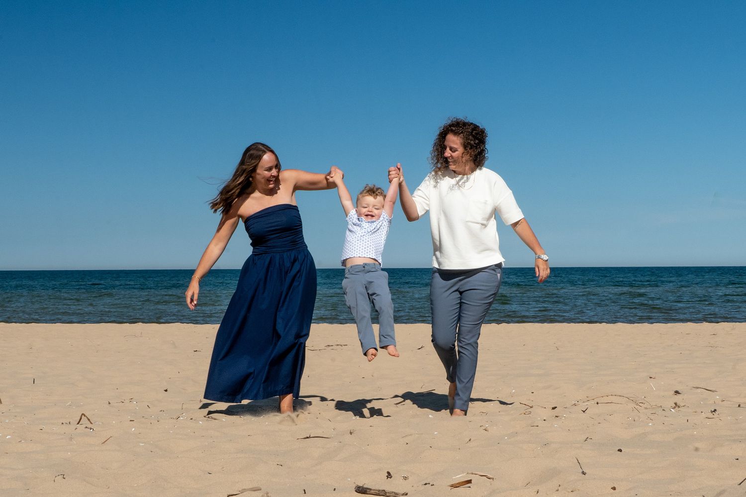 Family of three enjoying a sunny beach day, parents swinging toddler by hands on sandy shore with blue sky.