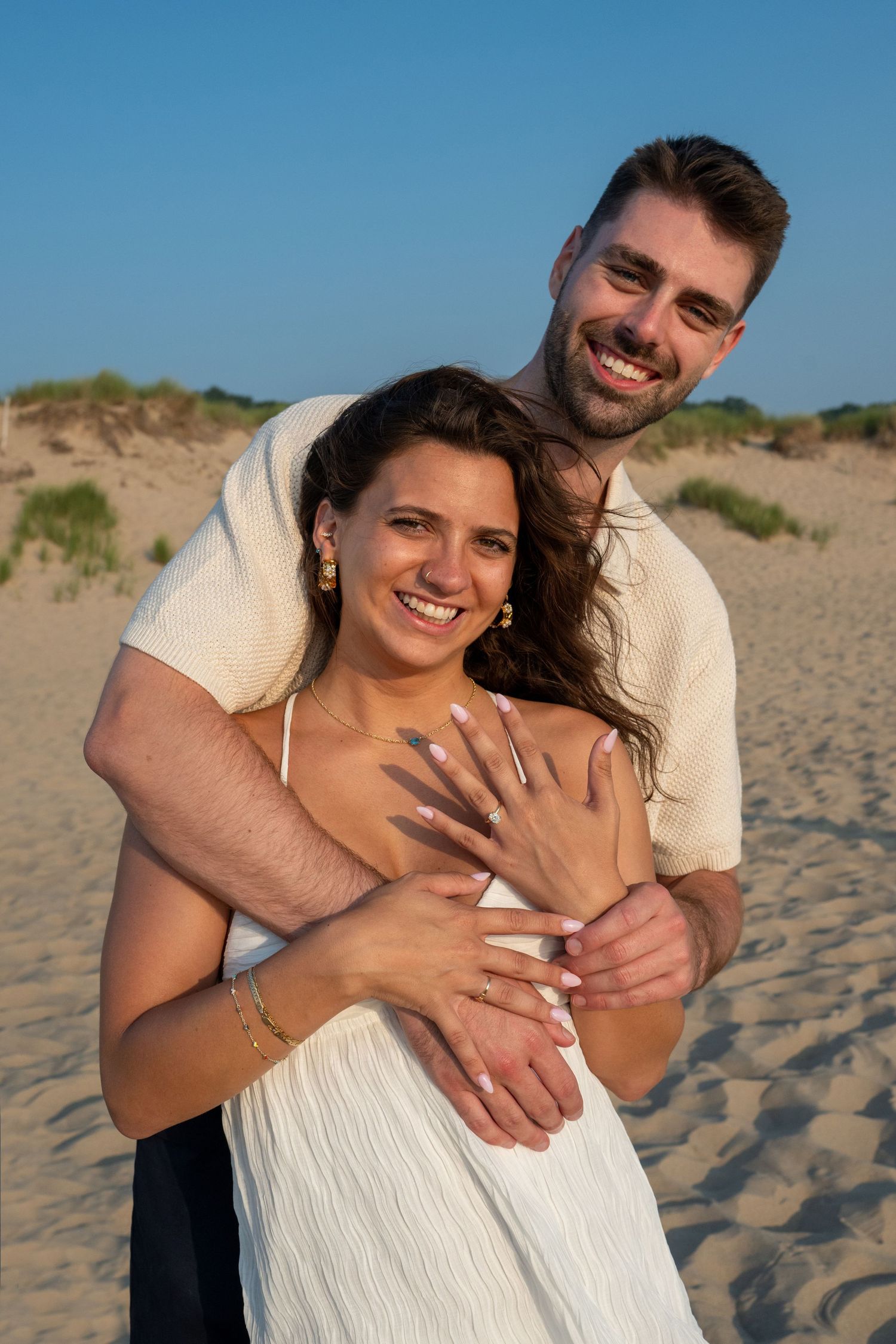 Happy couple embracing on sandy beach, woman showing engagement ring, smiling outdoors in golden hour light.