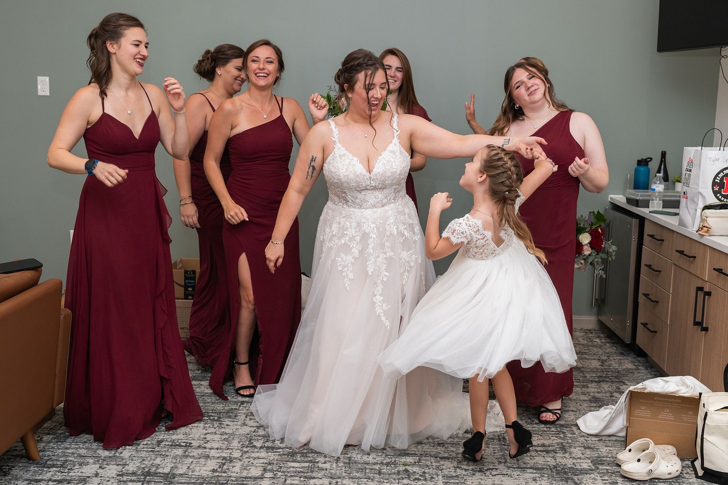 Bride in white lace gown laughs with bridesmaids in burgundy dresses and flower girl in white dress indoors.