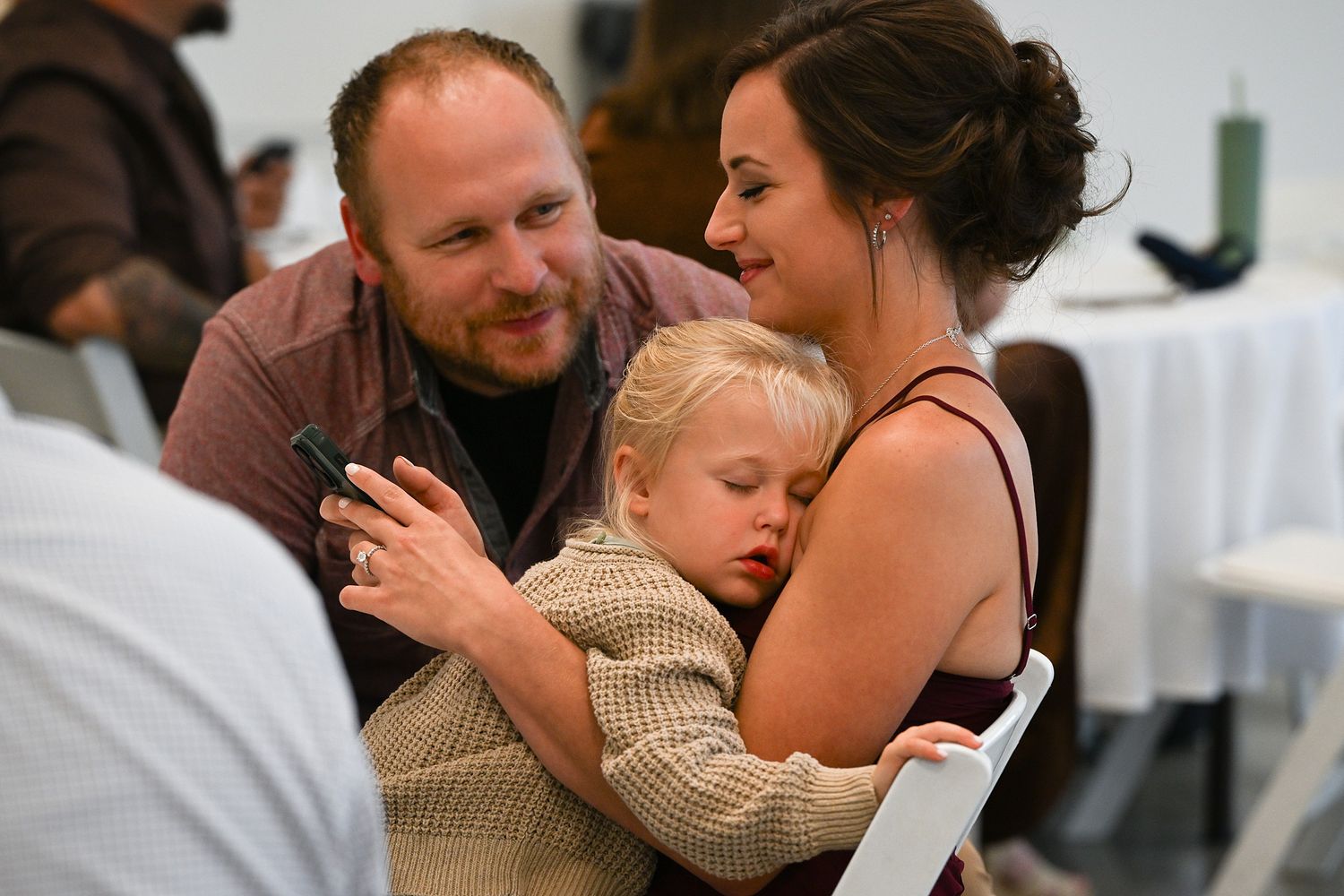 A couple tenderly holds their sleepy toddler during a wedding reception at a white-decorated venue.