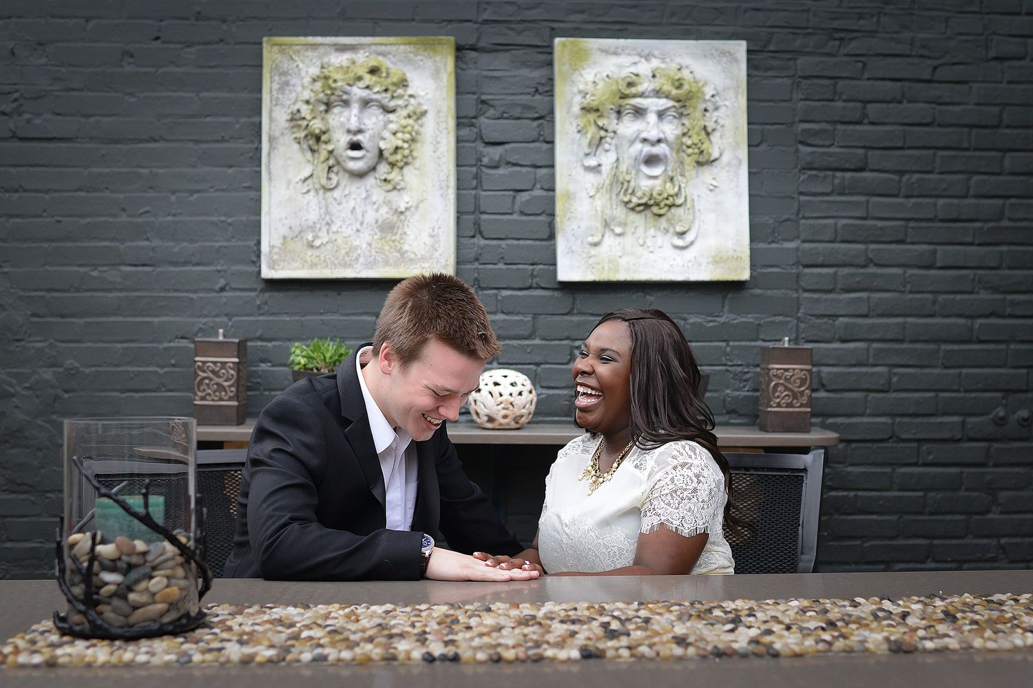 Interracial couple laughing together at outdoor table with decorative stone face wall art on dark brick background.