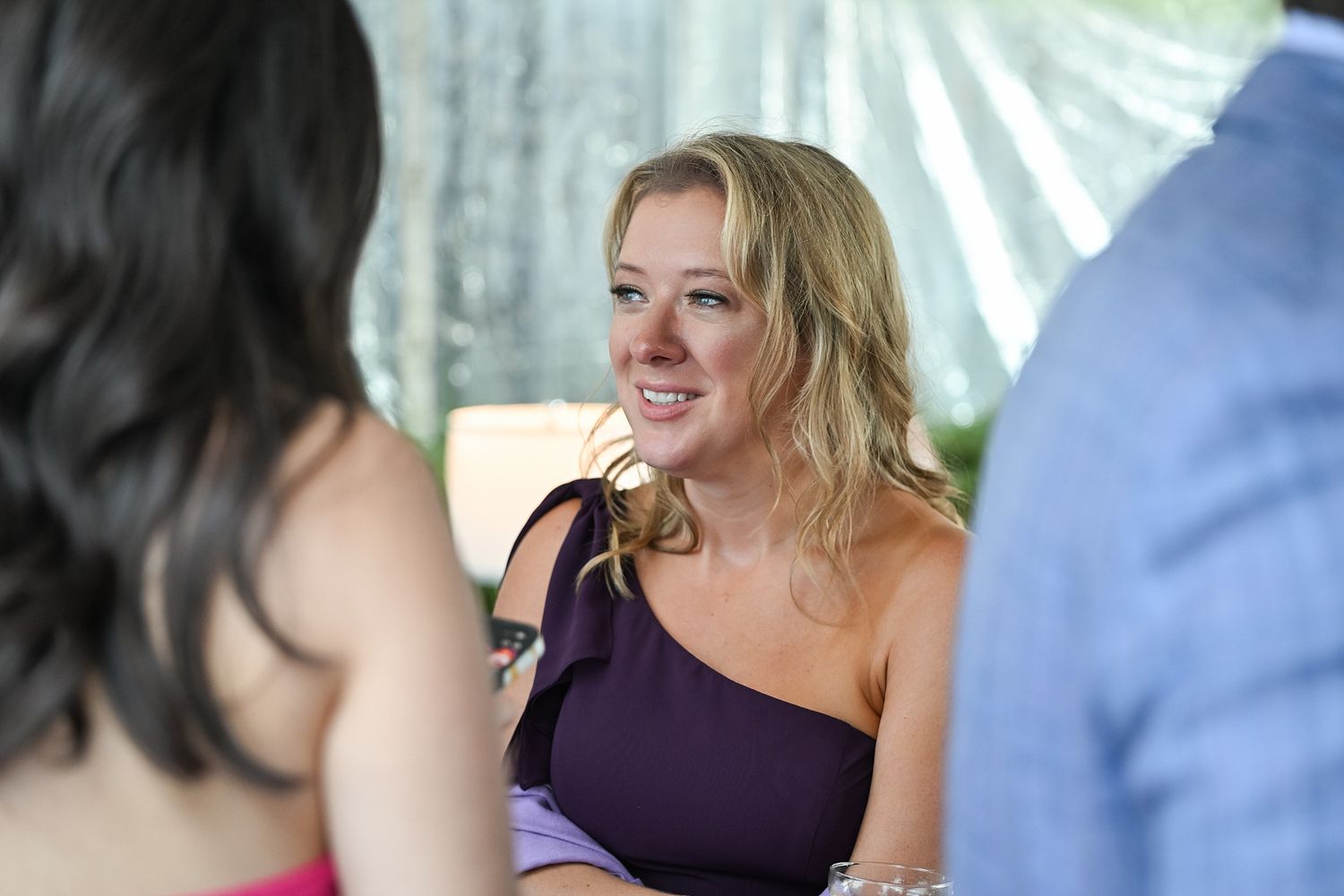 A person in a purple one-shoulder dress smiles while engaged in conversation at a social gathering.