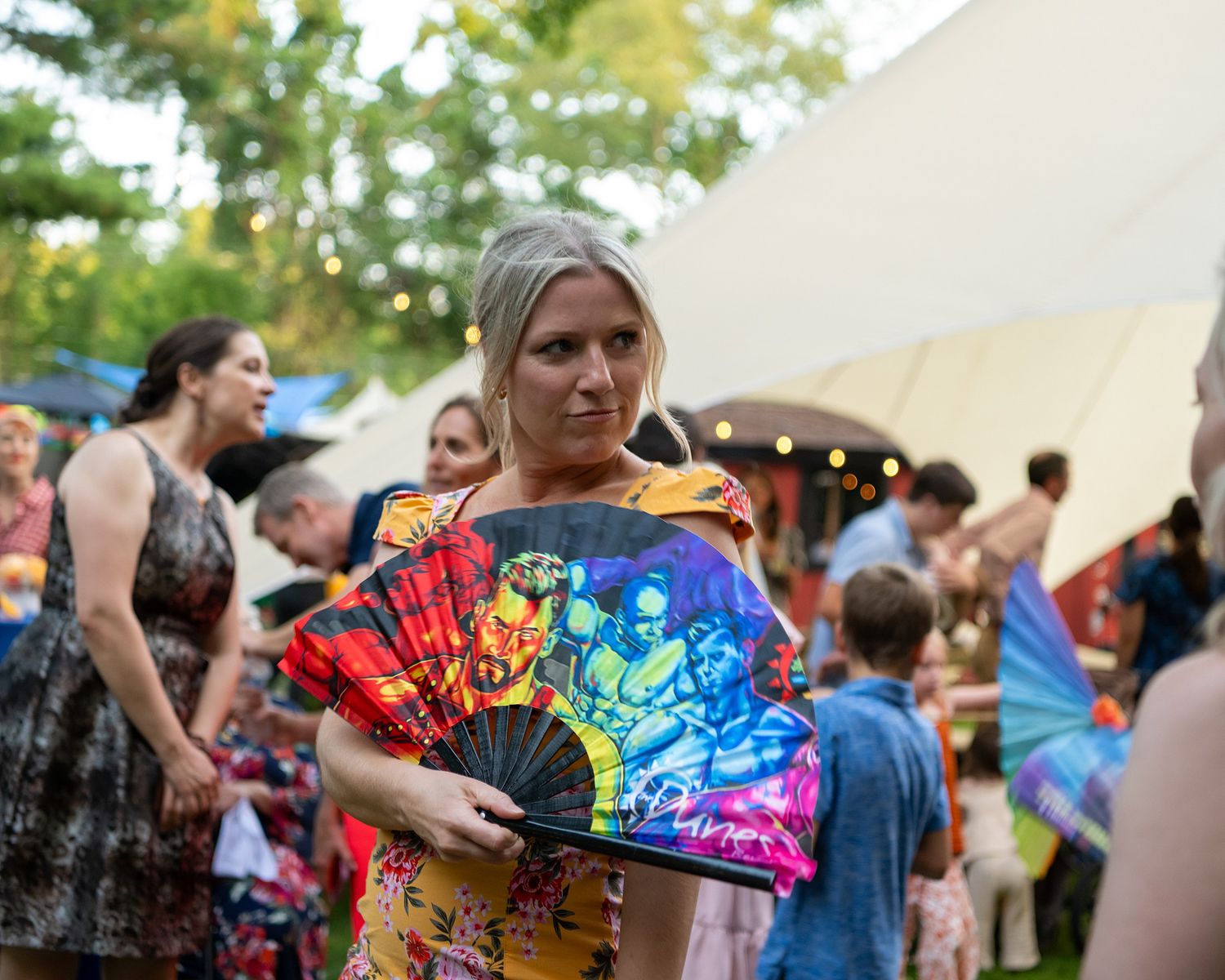 A festival attendee holds up a vibrant rainbow-colored folding fan against a backdrop of tents and people.