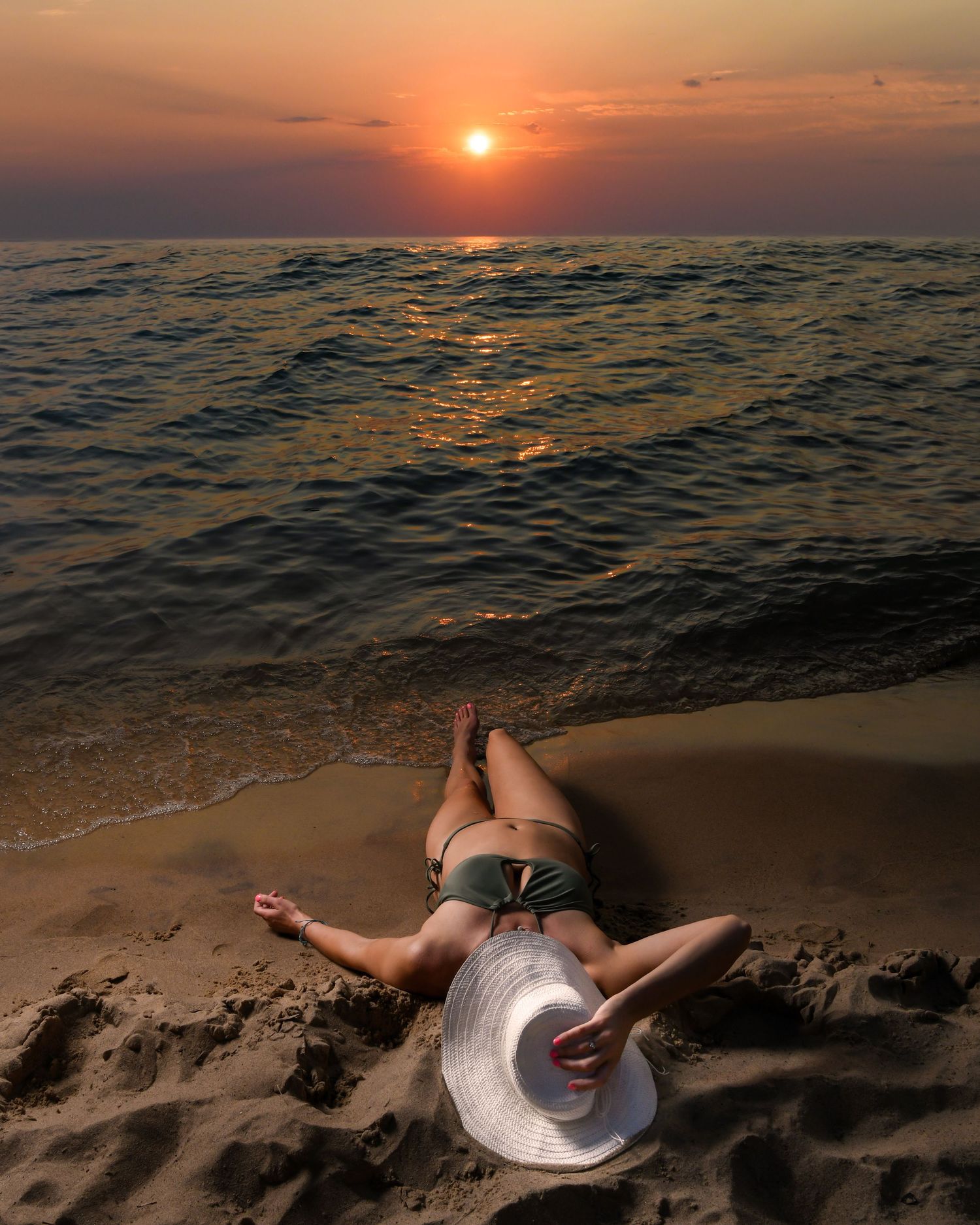 Person relaxing on beach with white hat during vibrant orange sunset over ocean waves.