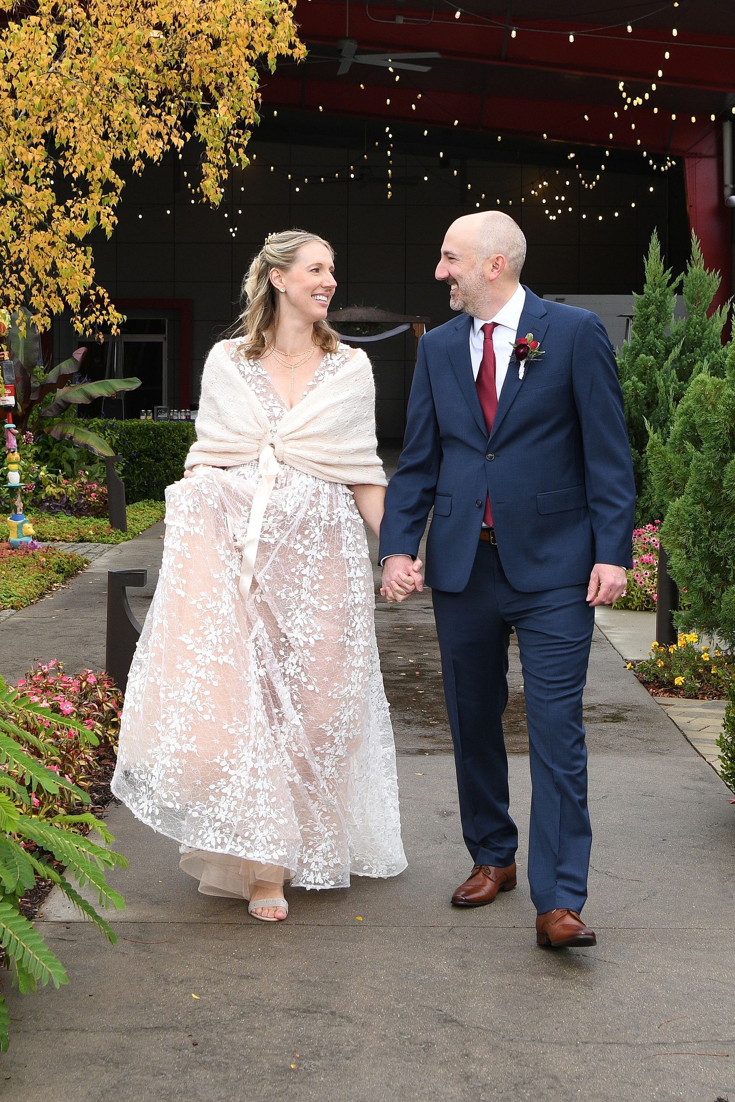 Couple walking together outdoors in formal attire with autumn foliage and string lights visible.
