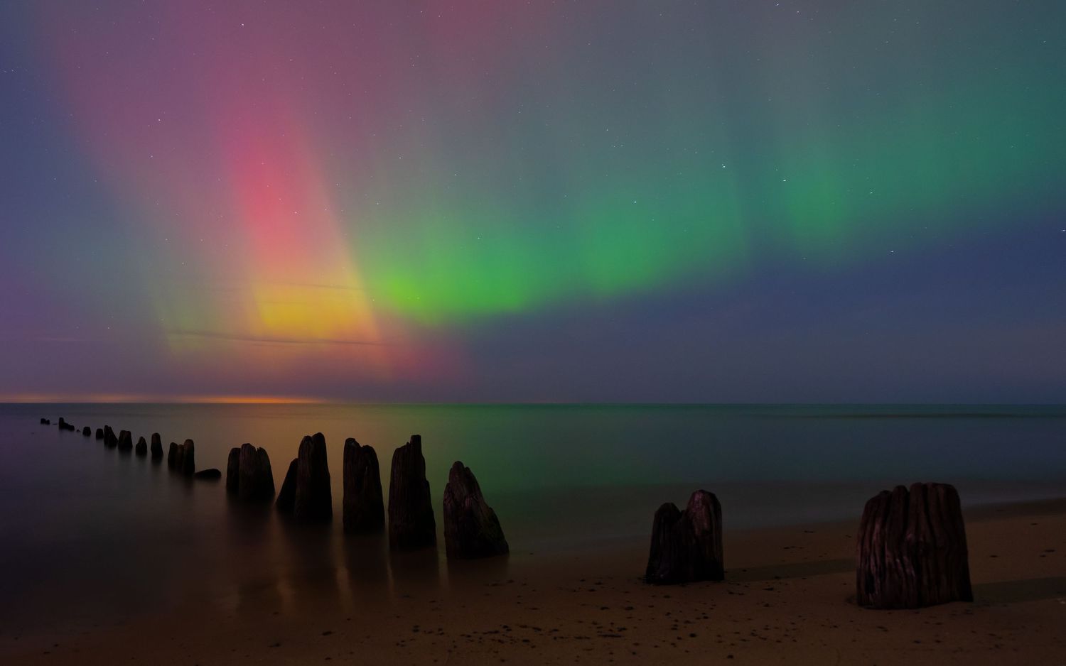 Aurora borealis illuminates the night sky with vibrant green and red hues over a calm sea with weathered wooden posts.