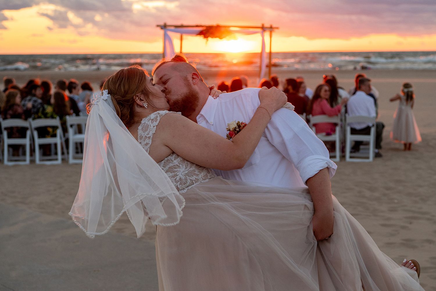Couple shares romantic kiss at sunset beach wedding ceremony with guests seated on white chairs in background.