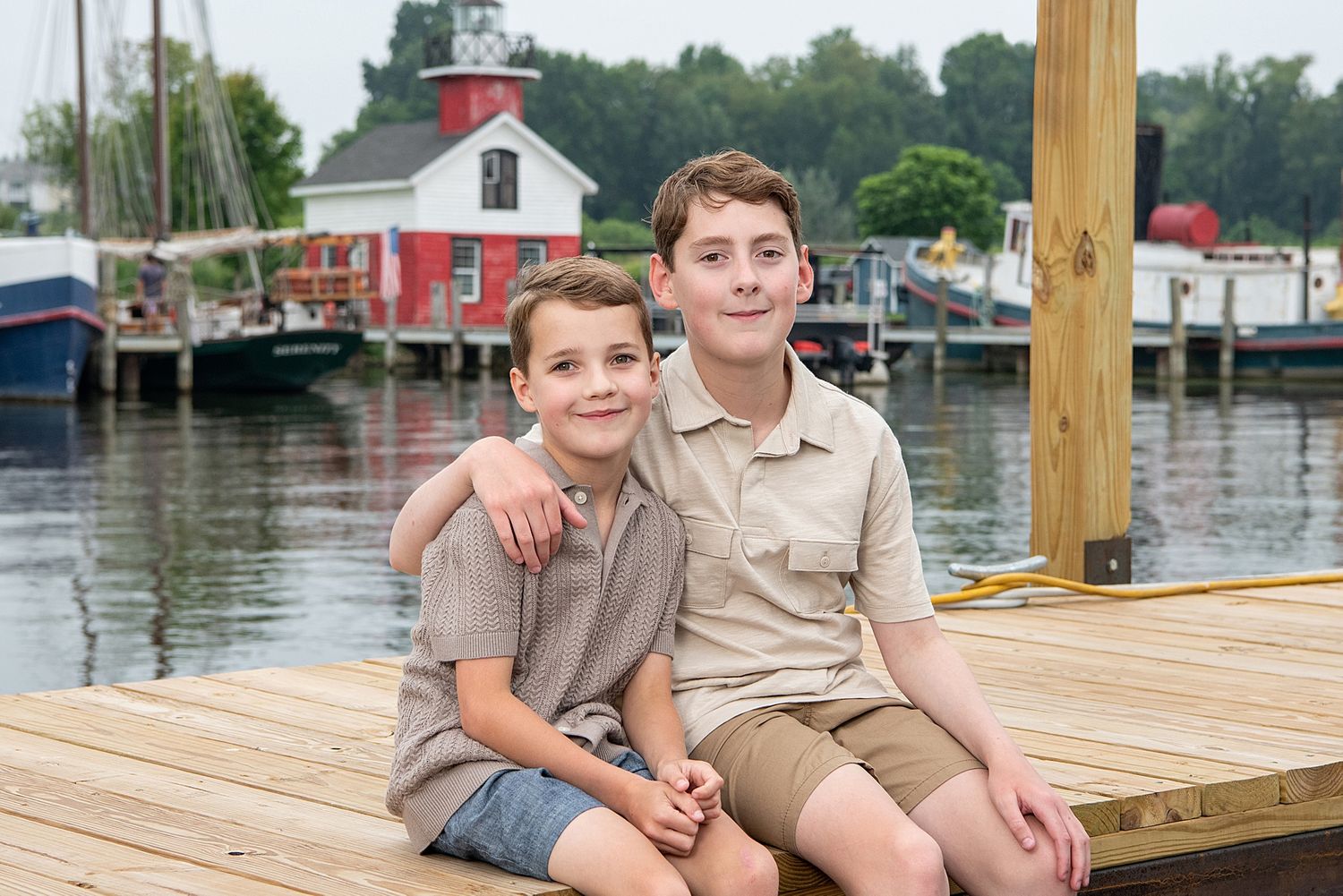 Two young people sit together on a wooden dock with a red boathouse visible in the background.