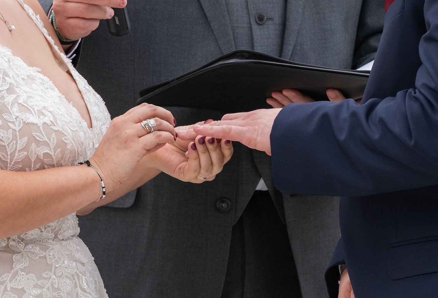 Bride places wedding ring on grooms finger during outdoor ceremony while officiant holds black book.