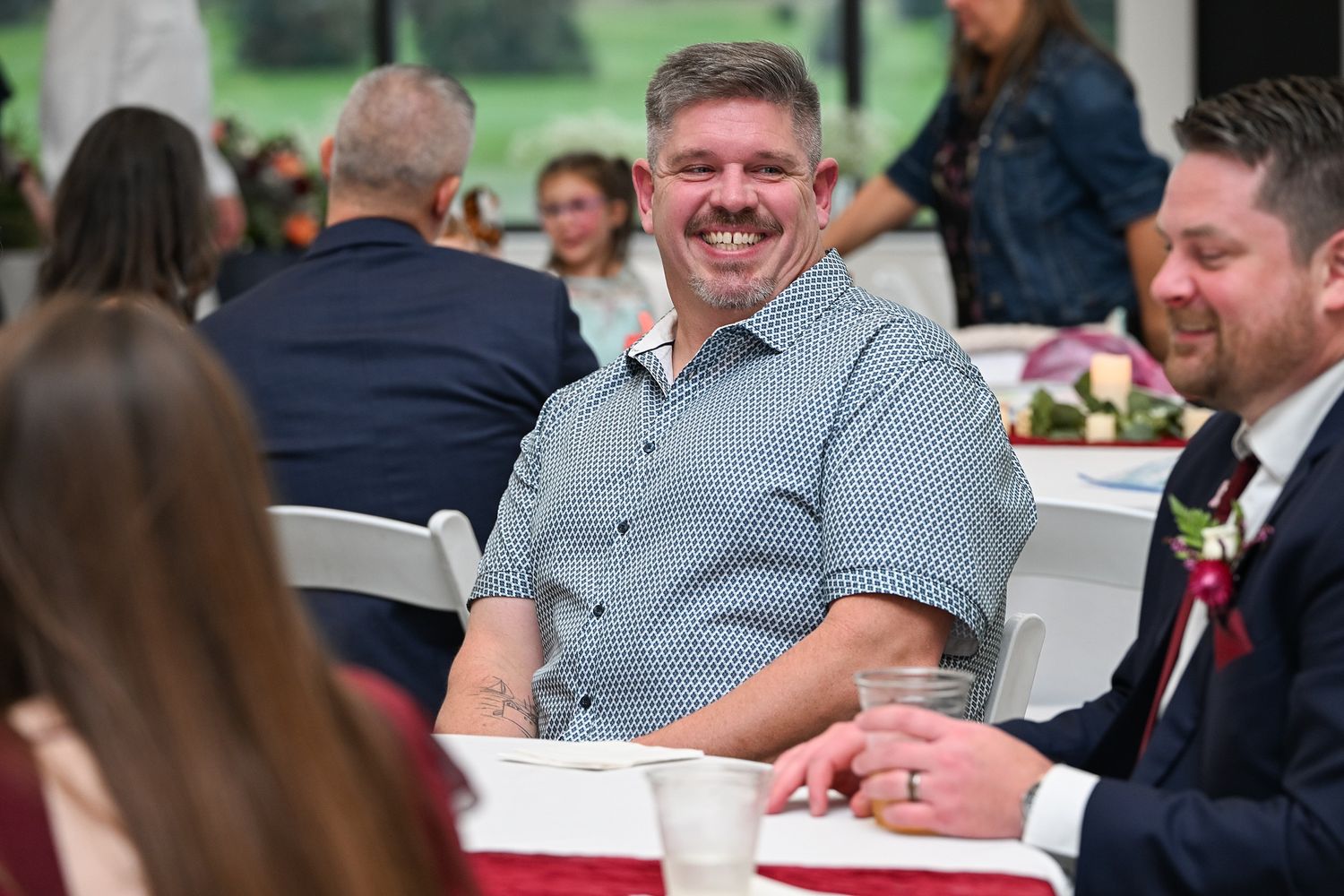Smiling man in blue shirt enjoying a wedding reception dinner, seated at a decorated table with guests.