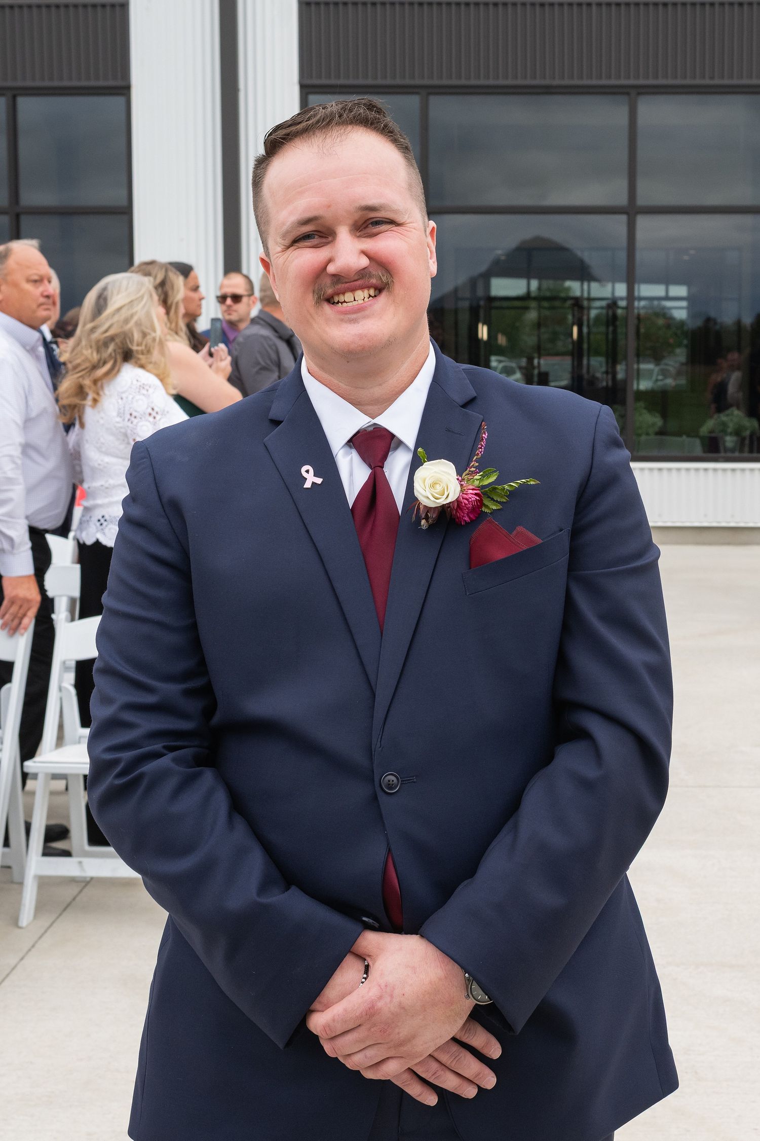 Smiling groom in navy blue suit with burgundy tie and floral boutonniere at outdoor wedding ceremony.