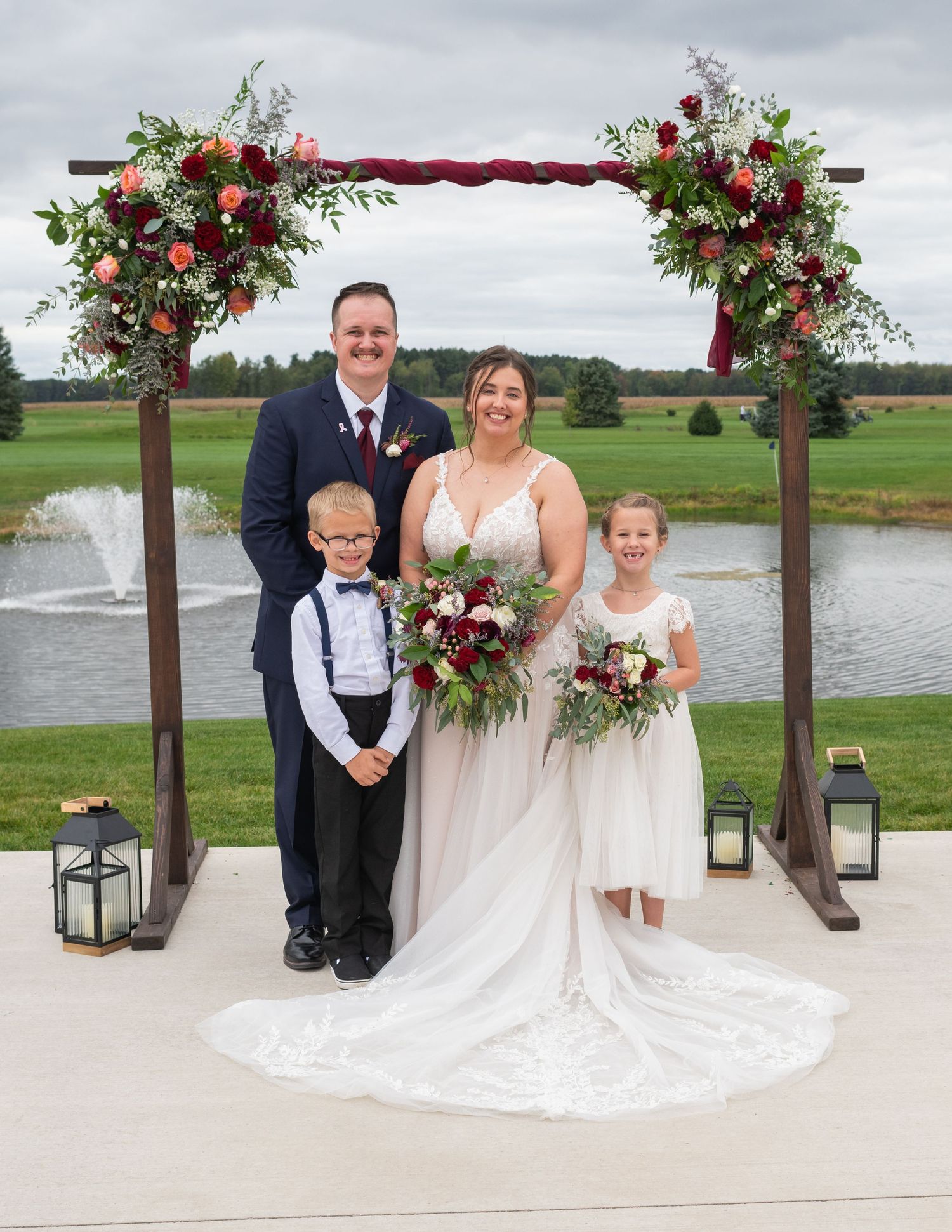 Wedding family portrait under floral arch with groom, bride, ring bearer, and flower girl by pond.