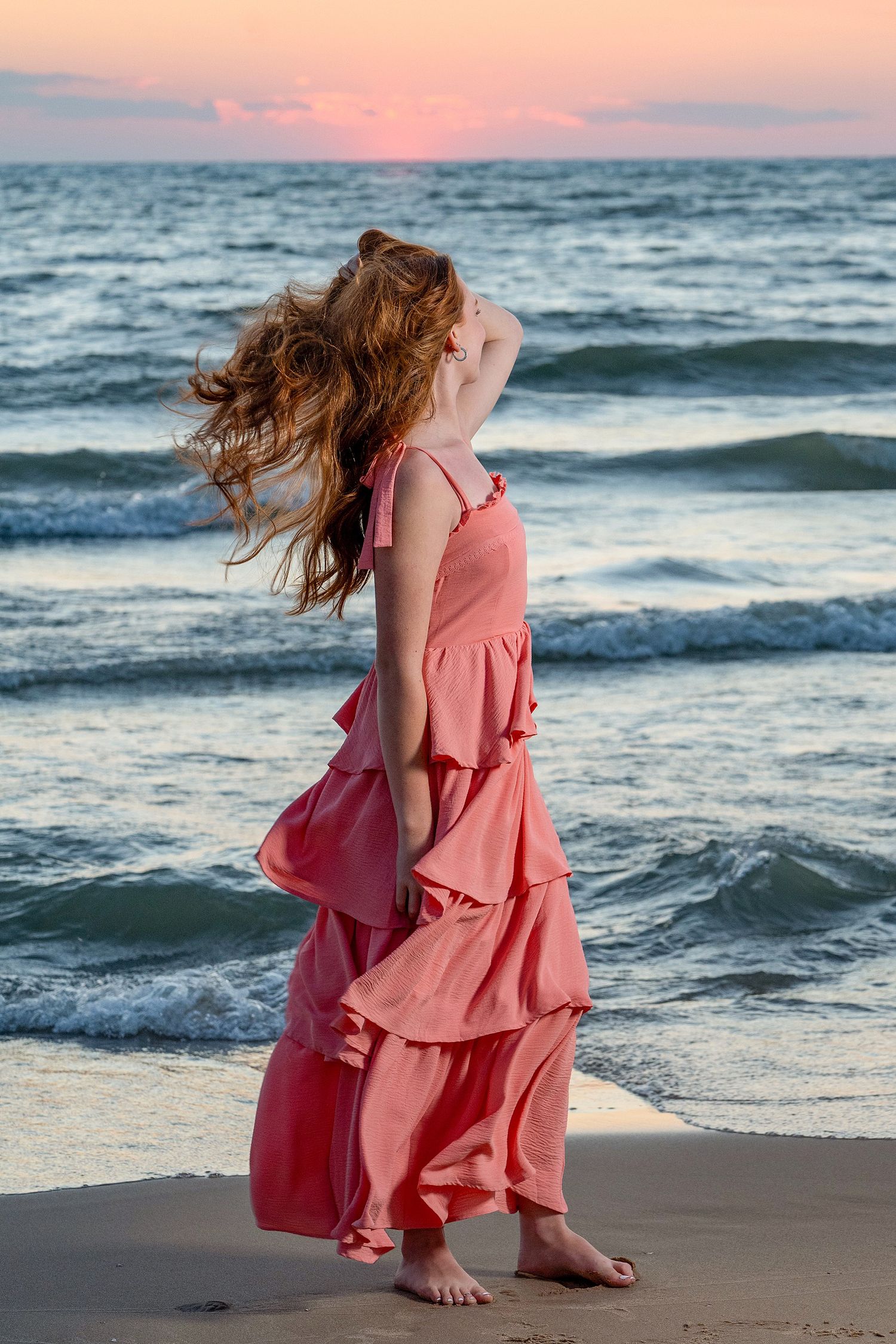 Young girl in pink ruffled maxi dress stands barefoot on beach at sunset, hair blowing in wind.