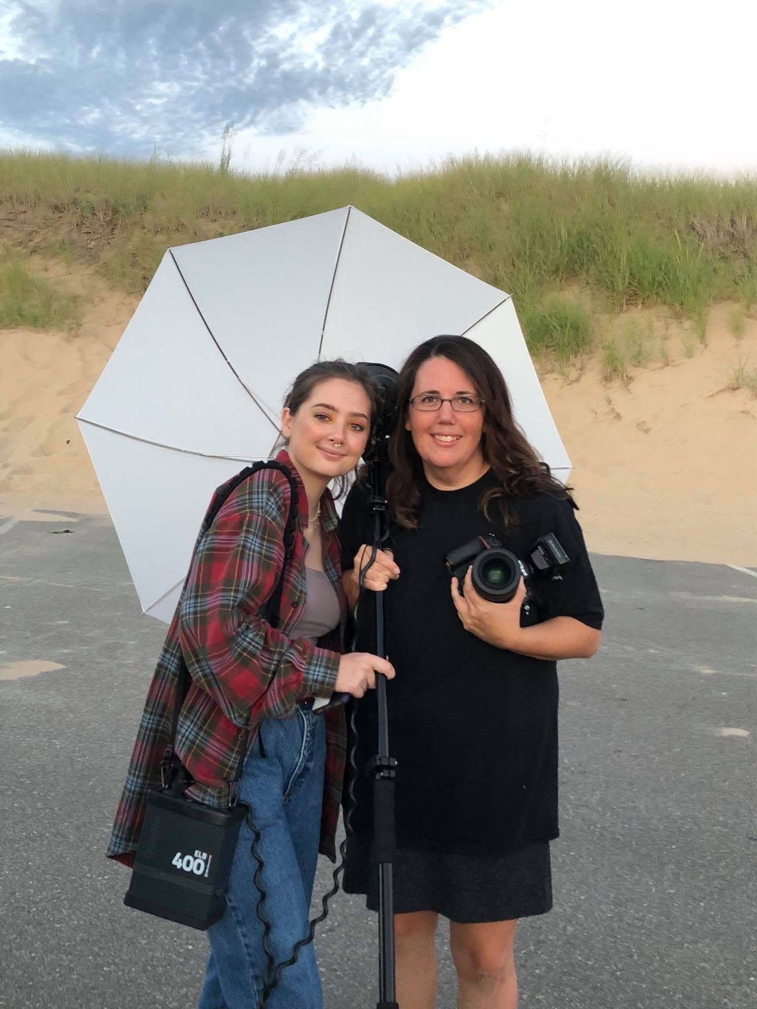 Photographers sharing a white umbrella while holding camera gear on a beach path with sand dunes.