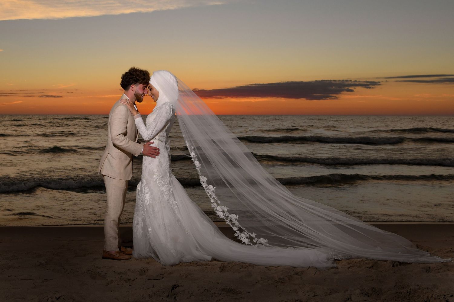 Bride and groom share a romantic kiss on the beach at sunset, with a flowing white veil and ocean waves behind them.