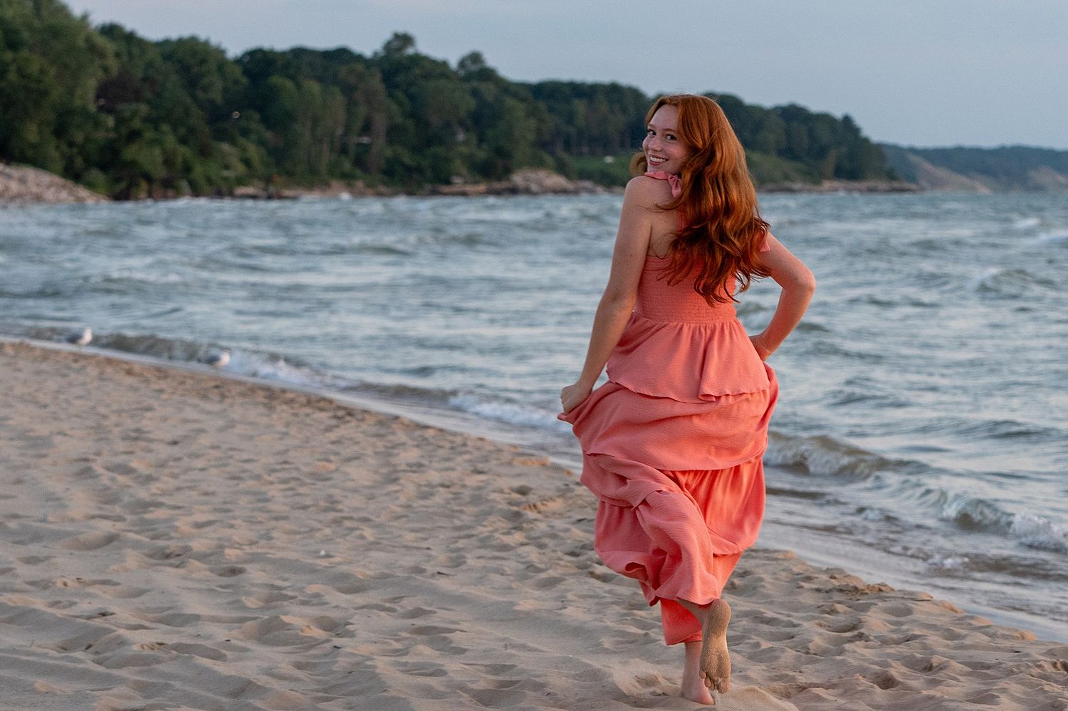 A smiling redhead woman in a coral dress walks along a sandy beach with waves and trees in the background.