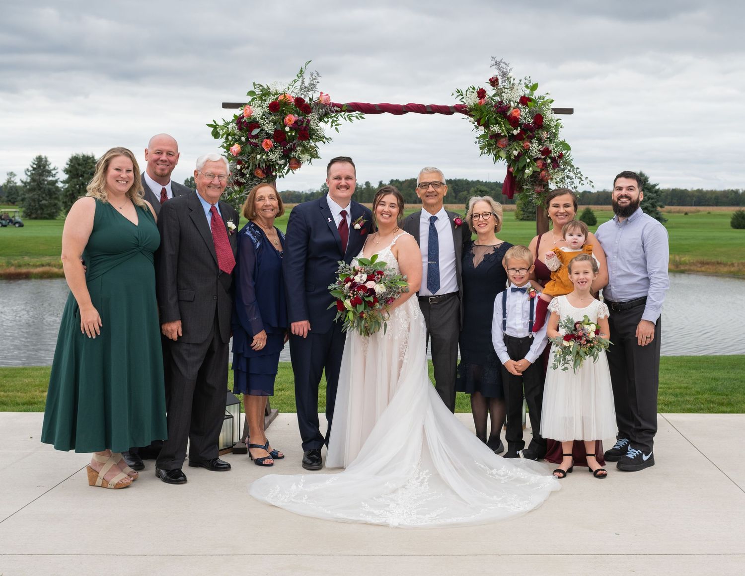 Wedding party poses outdoors under floral arch with bride and groom, overlooking scenic pond backdrop.