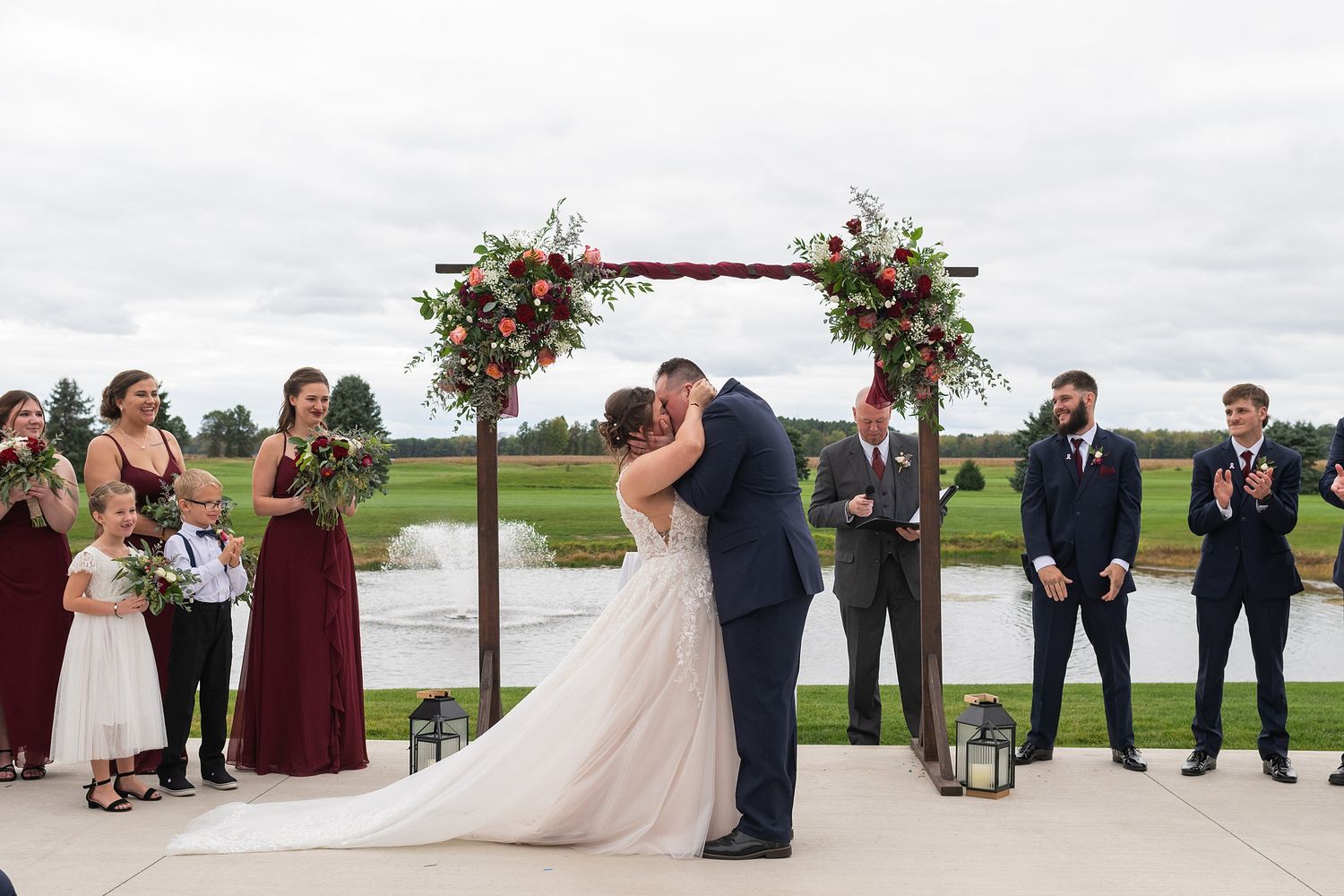 Bride and groom share first kiss under floral arch at outdoor wedding ceremony by a pond.