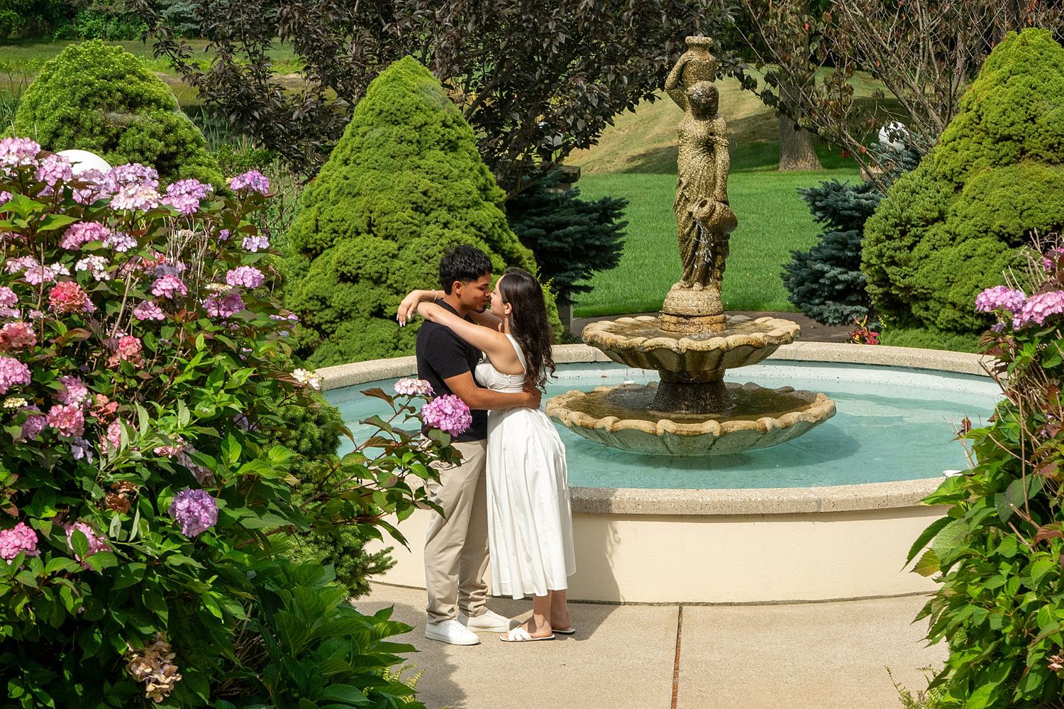 Couple embracing romantically by a stone tiered fountain in a lush garden with pink hydrangeas.