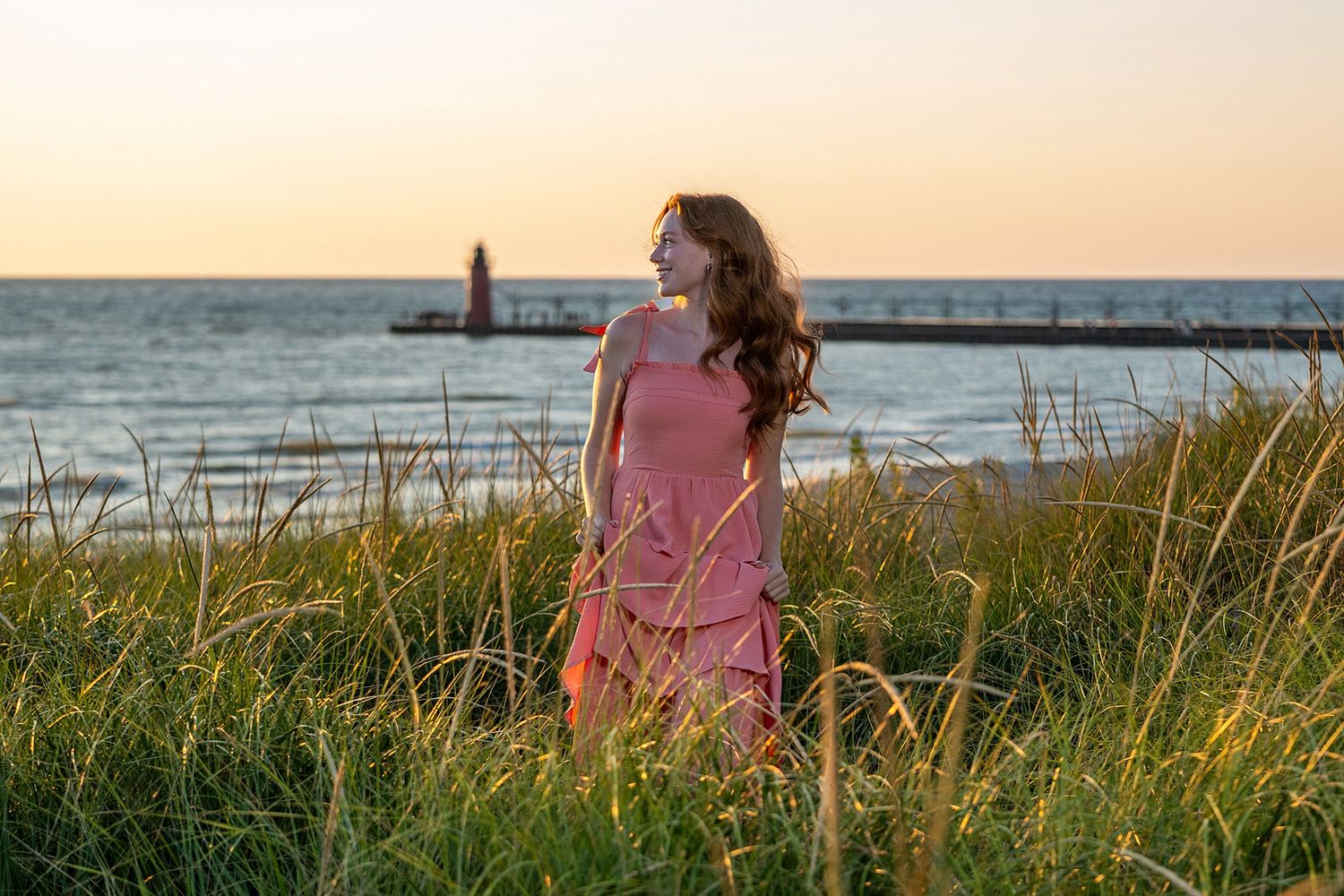 Woman in pink dress standing in tall grass at sunset near a lighthouse and calm ocean waters.