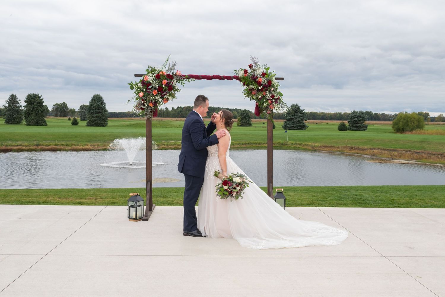 Bride and groom share first kiss under floral arch at outdoor wedding venue beside a serene pond.