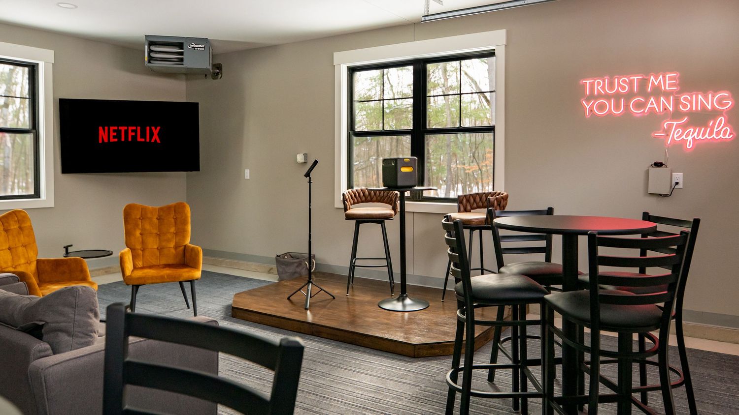 Modern entertainment room with Netflix TV, neon Tequila sign, bar tables, velvet chairs, and karaoke microphone stand.