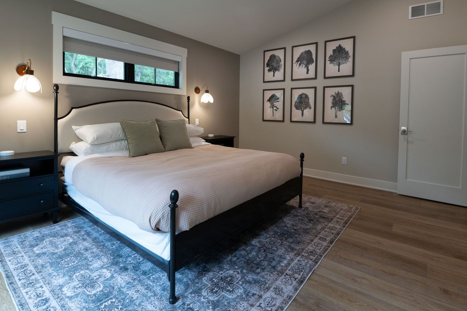 Modern farmhouse bedroom with black metal bed frame, blue area rug, hardwood floors, and botanical wall art gallery.