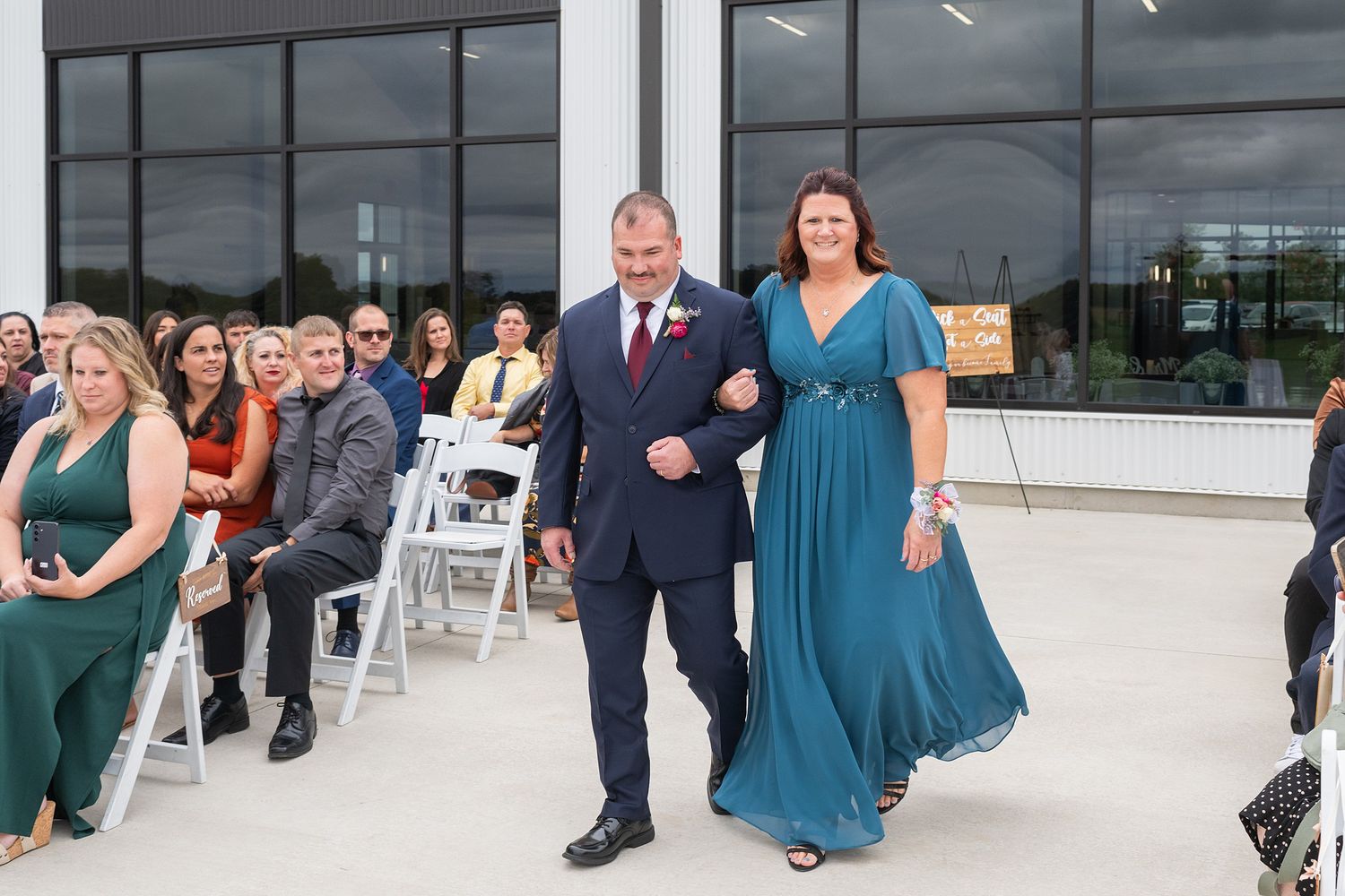 A man in a navy suit escorts a woman in a teal gown down the wedding aisle past seated guests.