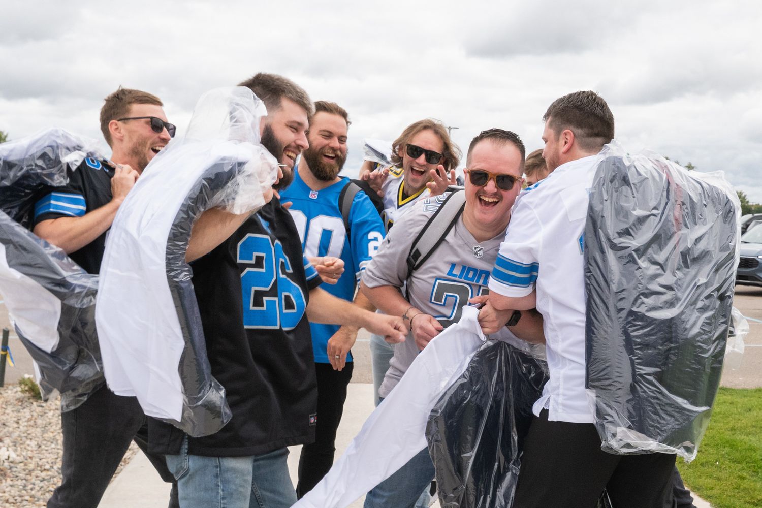 Detroit Lions NFL fans laughing and celebrating outdoors wearing team jerseys and rain ponchos at a game day event.